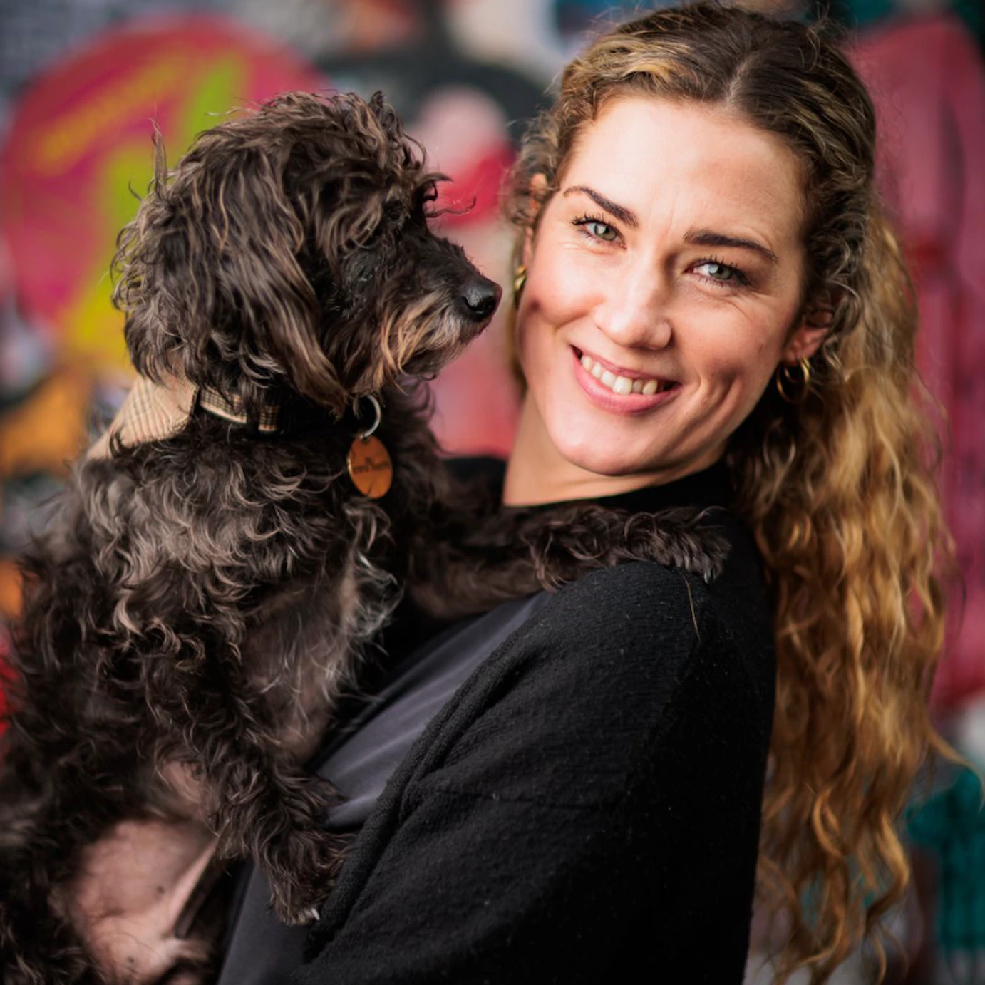 Kat Brown smiling to camera and holding a small black dog in her arms. In the background is a blurred out graffiti wall.