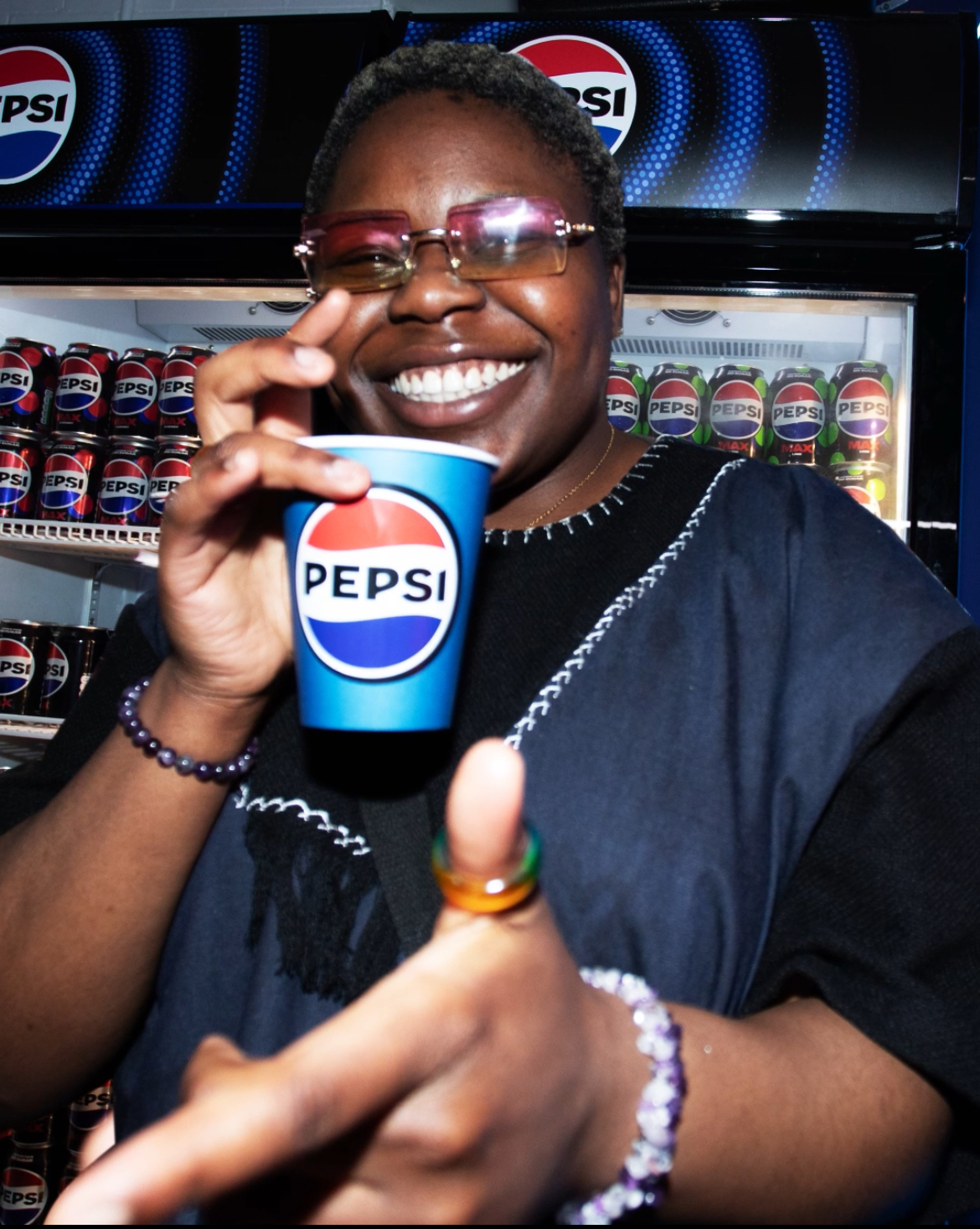 Baz smiling to camera in front of a fridge full of Pepsi cans. She is also holding a Pepsi paper cup.