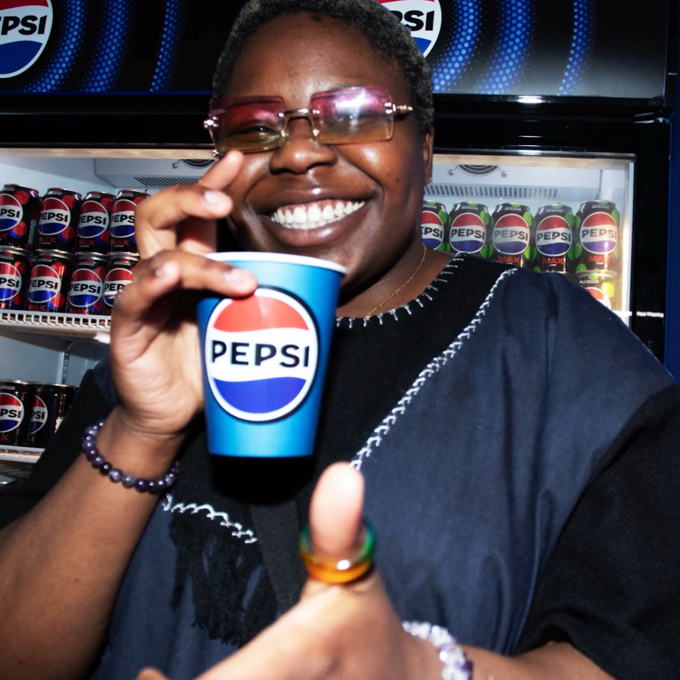 Baz smiling to camera in front of a fridge full of Pepsi cans. She is also holding a Pepsi paper cup.