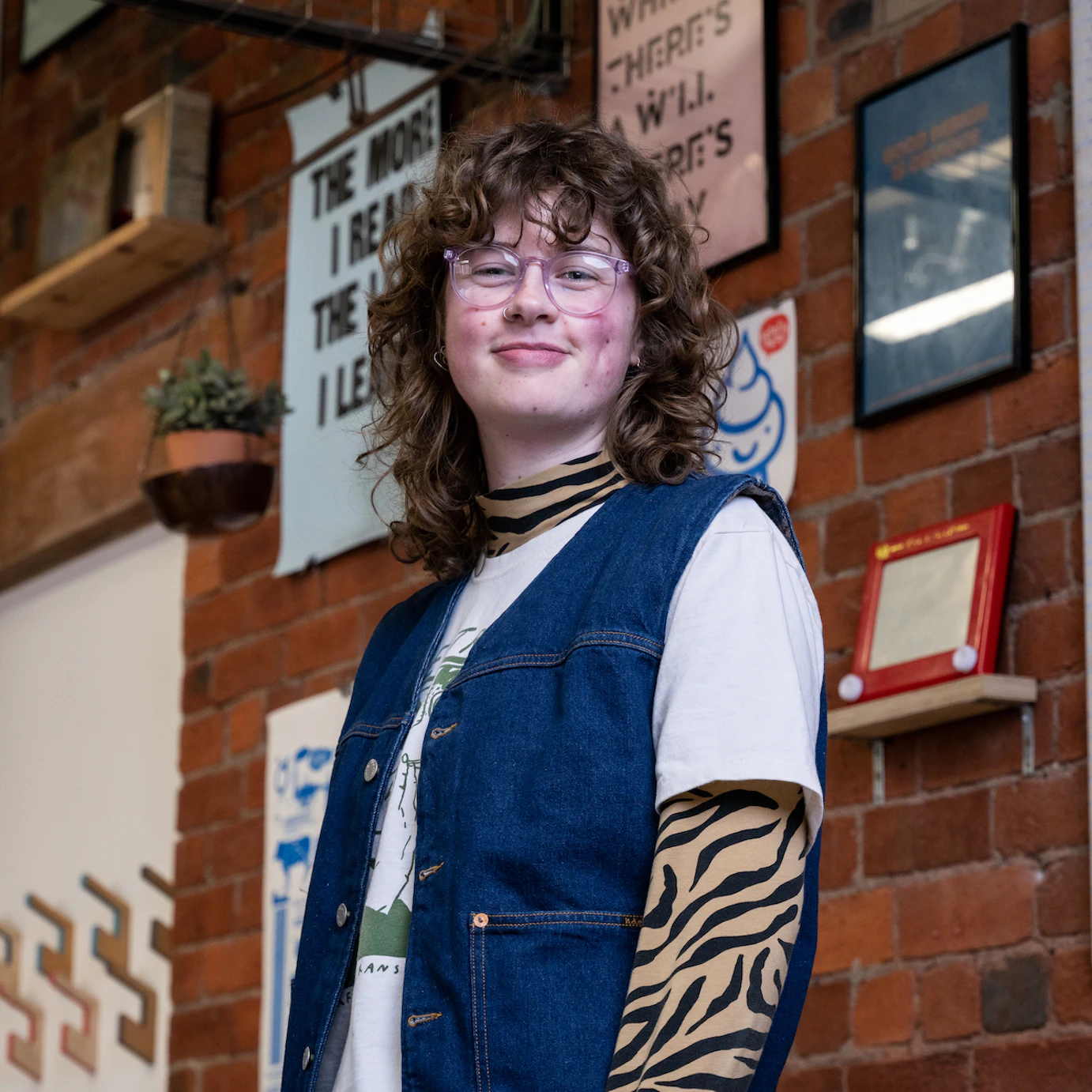Claire Hamilton stood smiling to camera on a background of various frames hung on an indoor brick wall.