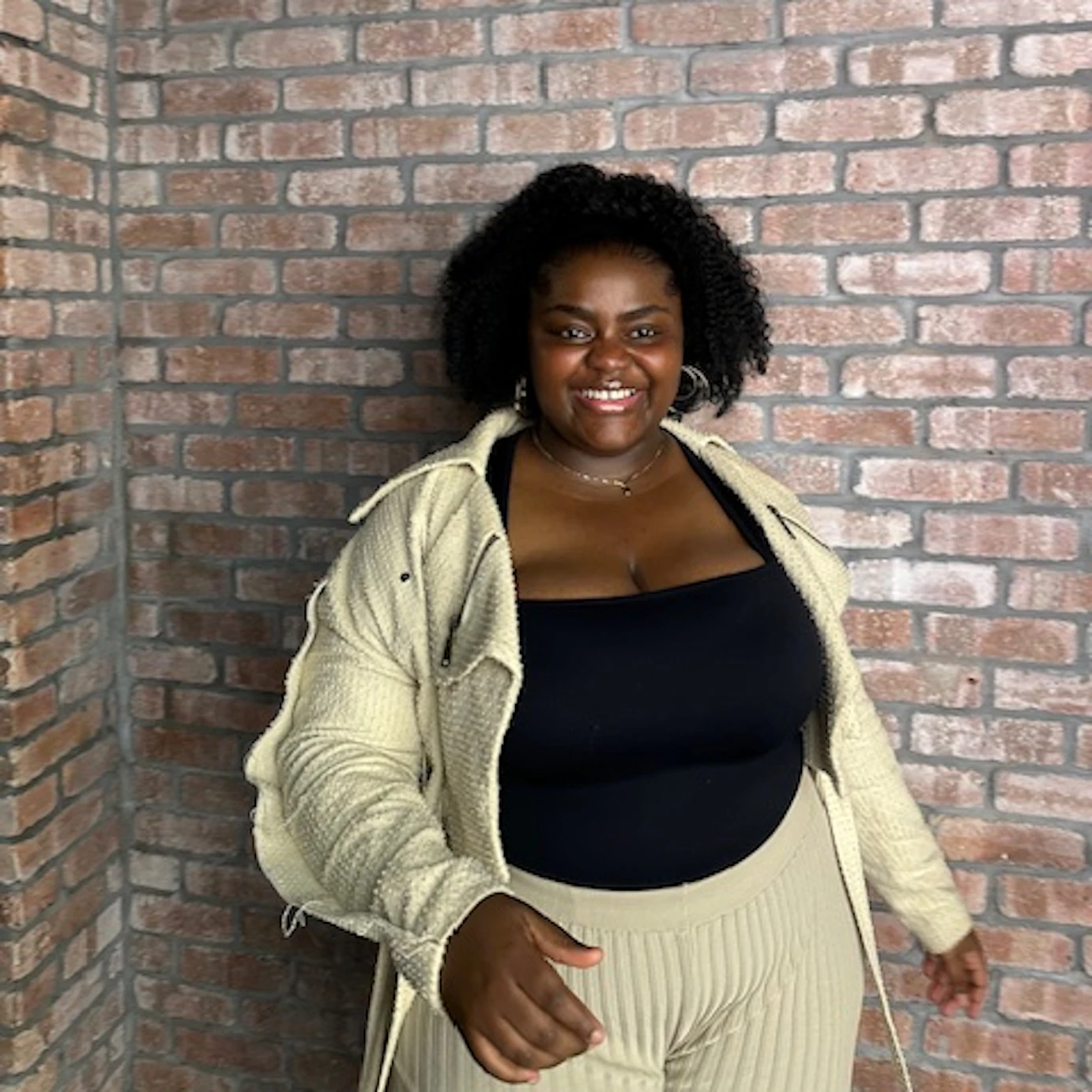 Tilly Tillz stood in the corner of a brick wall smiling just off camera.
