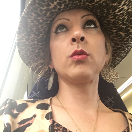 A selfie of Donna looking above the camera and pouting. Her large hat, earrings, blazer and top are all printed in leopard patterns.