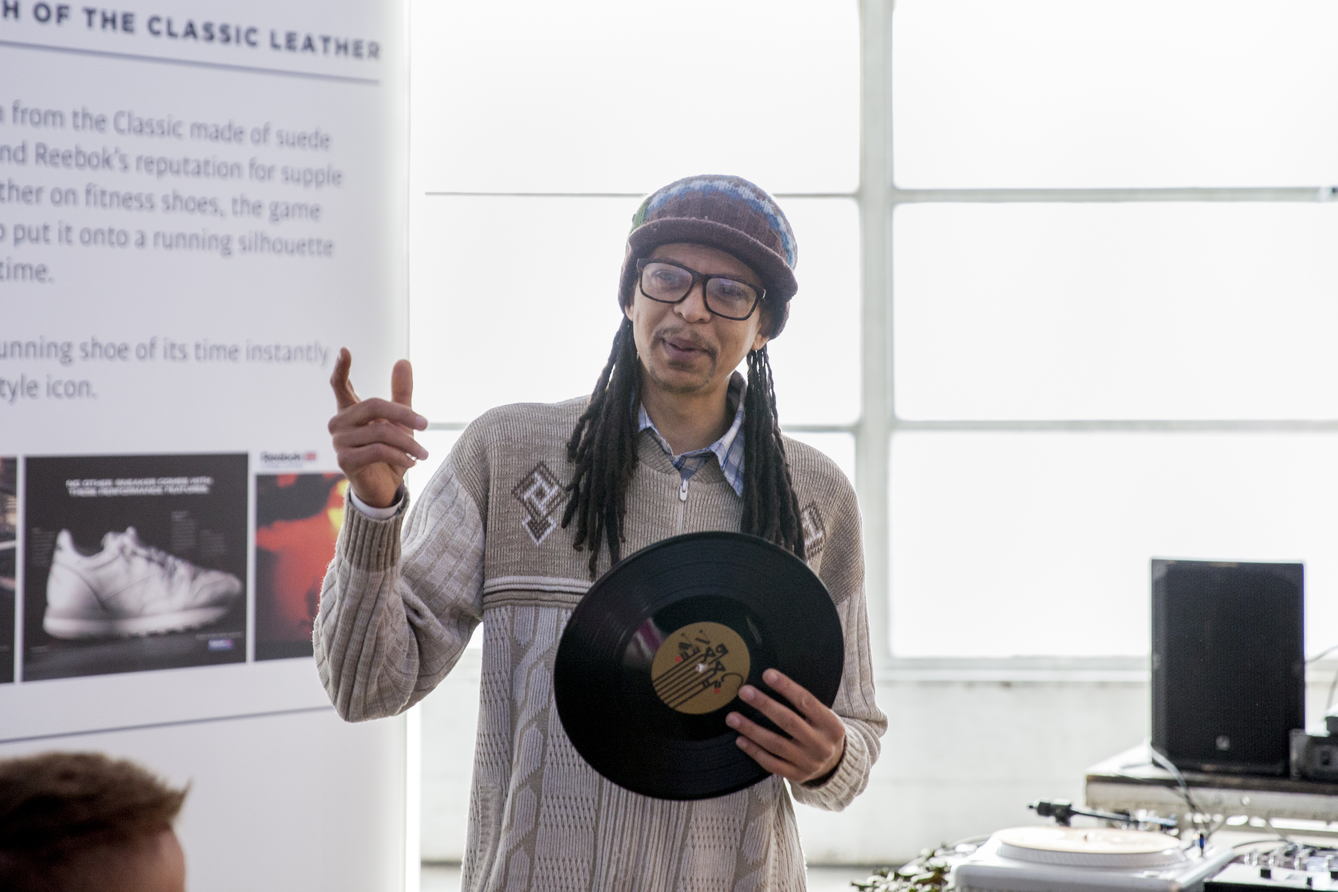 A person speaks to a crowd of people, holding a vinyl record with a set of DJ decks behind them.