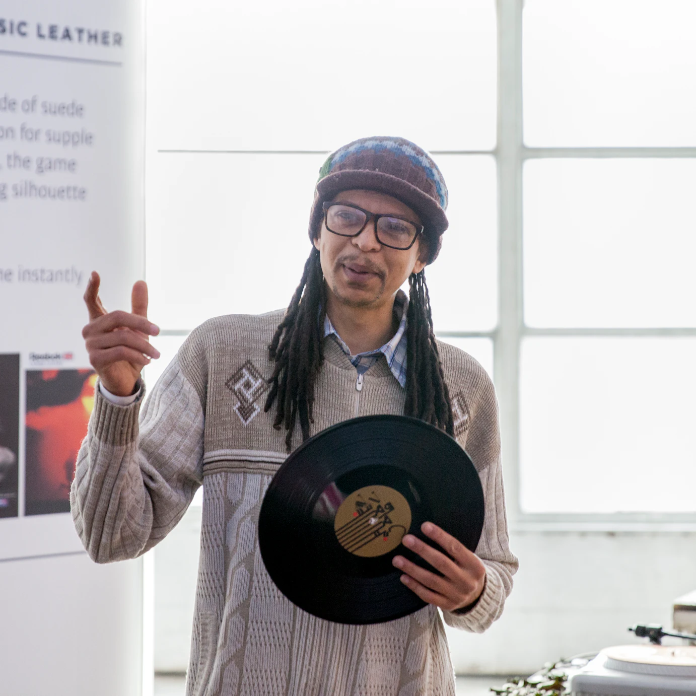 A person speaks to a crowd of people, holding a vinyl record with a set of DJ decks behind them.