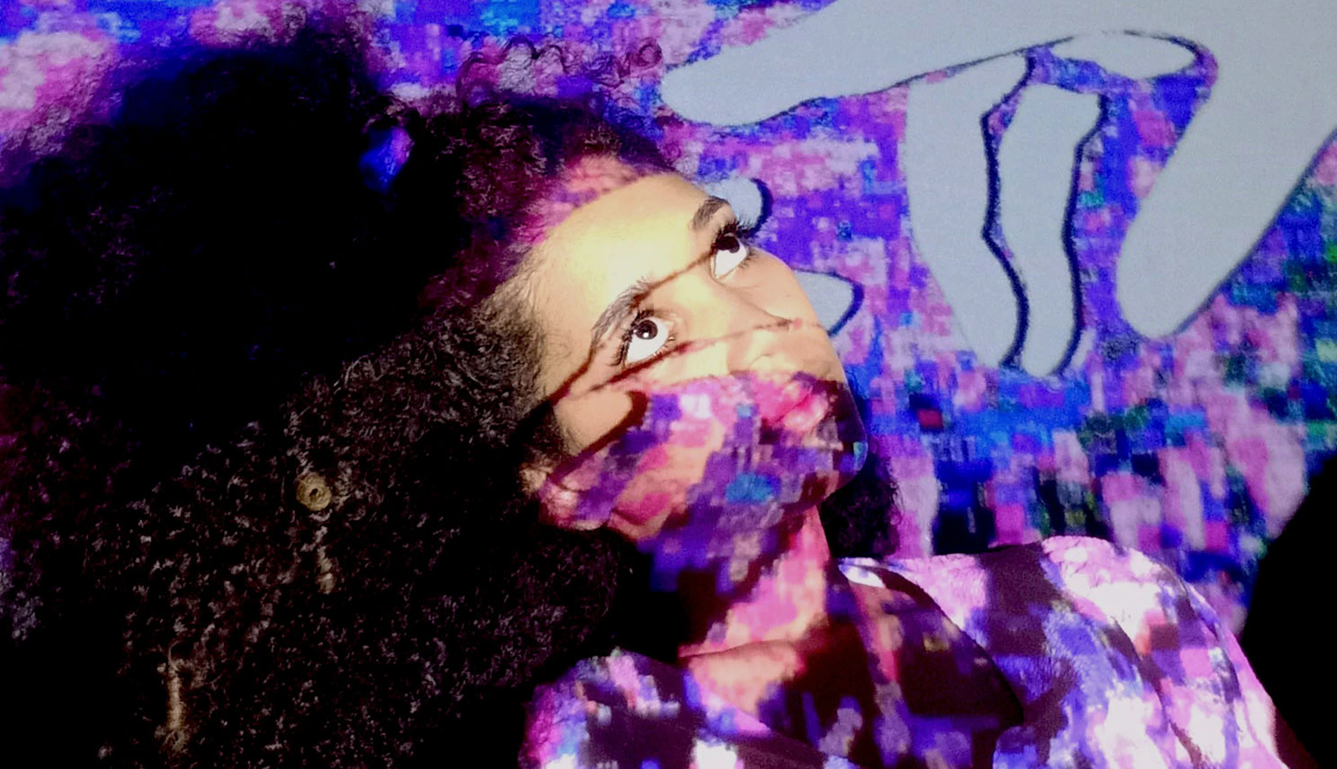 Alexis Maxwell looking up as a blue, pink and purple distorted and pixelated image is projected onto her. Also part of the projection is a white hand pointing towards Alexis's head which is tilted slightly as if anticipating being touched. Above the hand reads 'I just need a hug'.