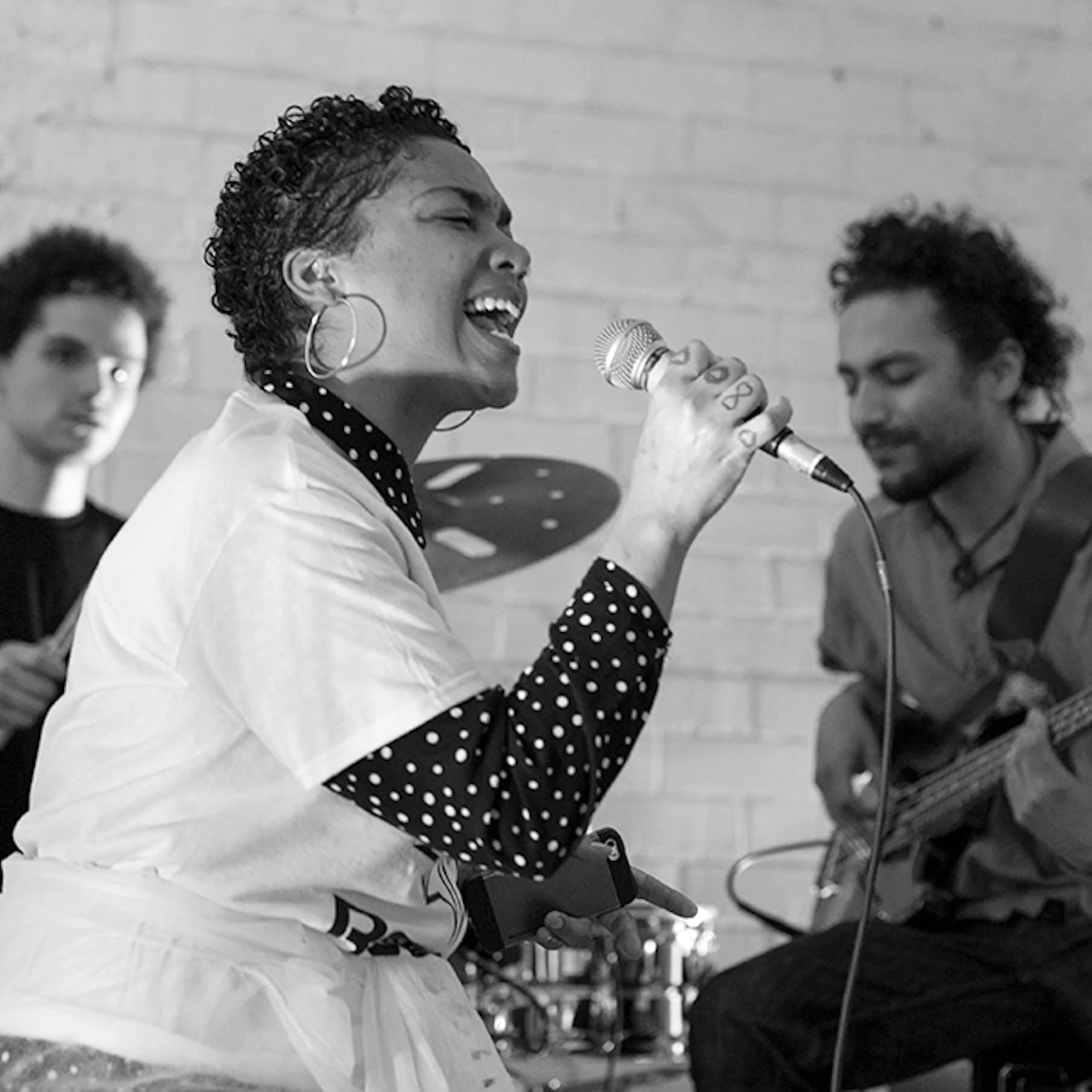 A black and white image of three musicians performing live together. In the background there is a drummer and a bassist. In the foreground, a vocalist sings into a microphone with their eyes closed.