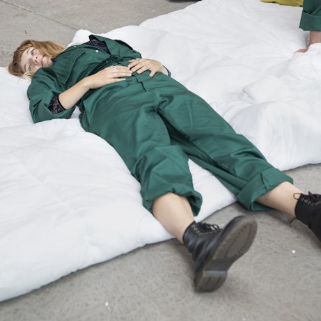 A residency artist dressed in a dark green boiler suit and black boots lay on a quilt on the floor and smiling.