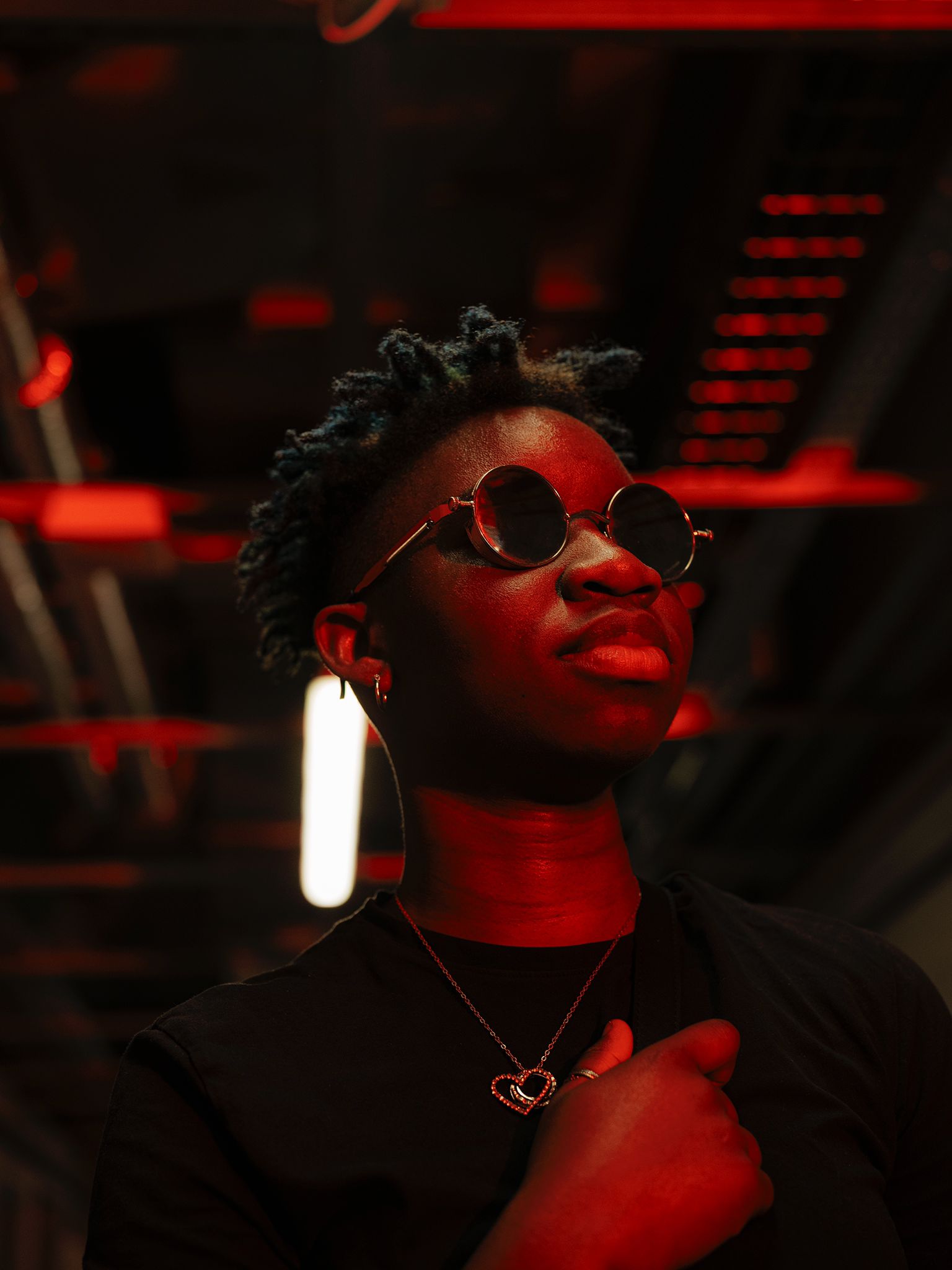Mara Cruz stood in a dark indoor space looking towards something with circular sunglasses on. The space and Mara have a red shadow illuminated from somewhere off camera.