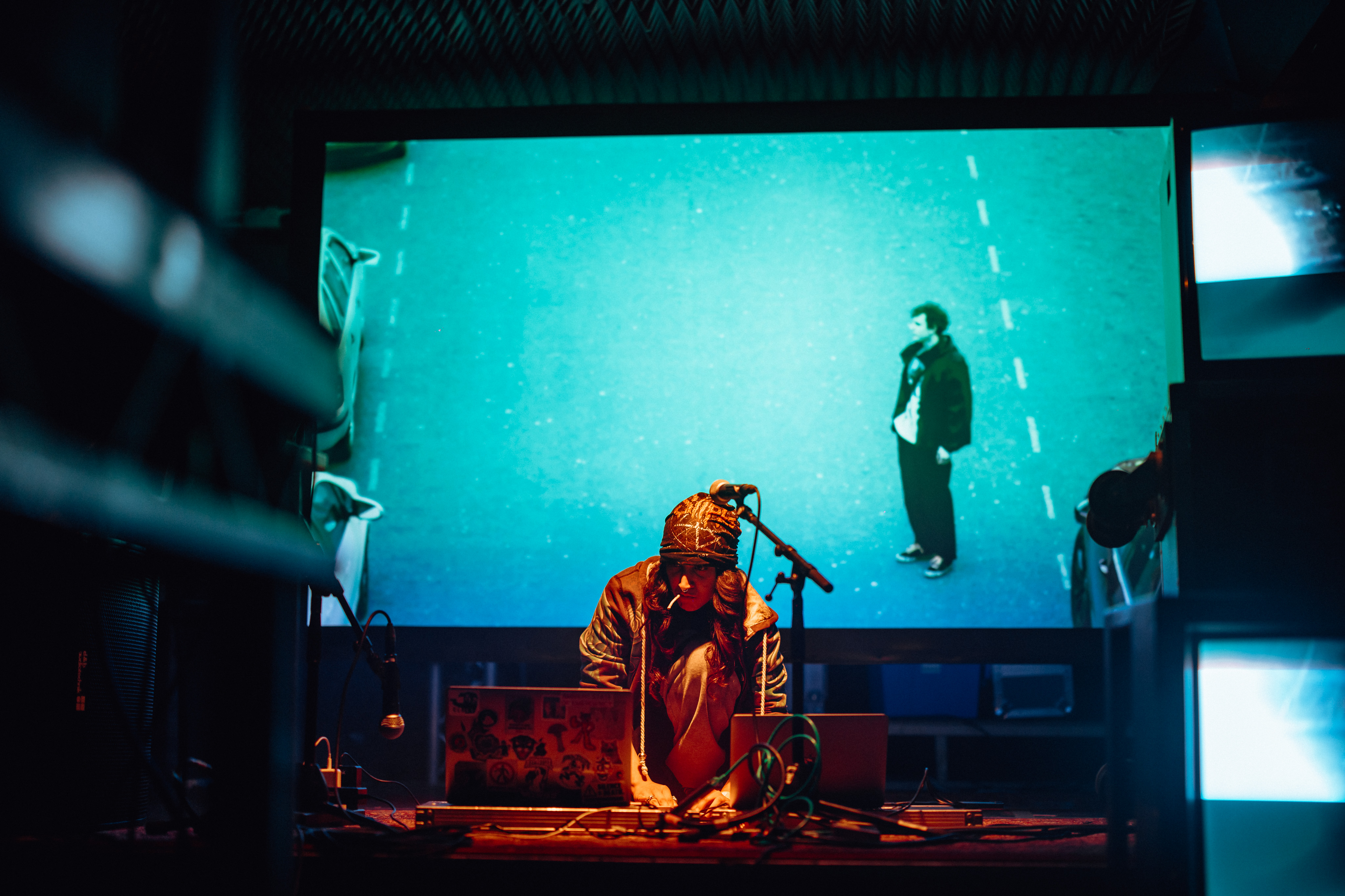 A musician kneels on the floor of the stage in front of a laptop. Behind them is a big screen projecting visuals which include an image of a man.