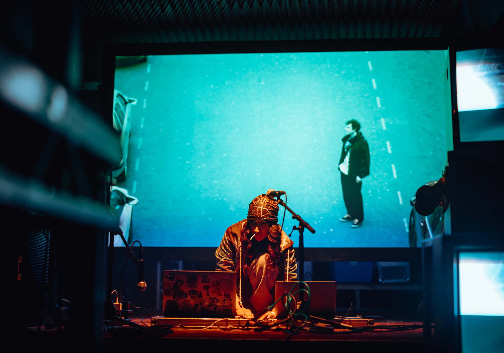 A musician kneels on the floor of the stage in front of a laptop. Behind them is a big screen projecting visuals which include an image of a man.