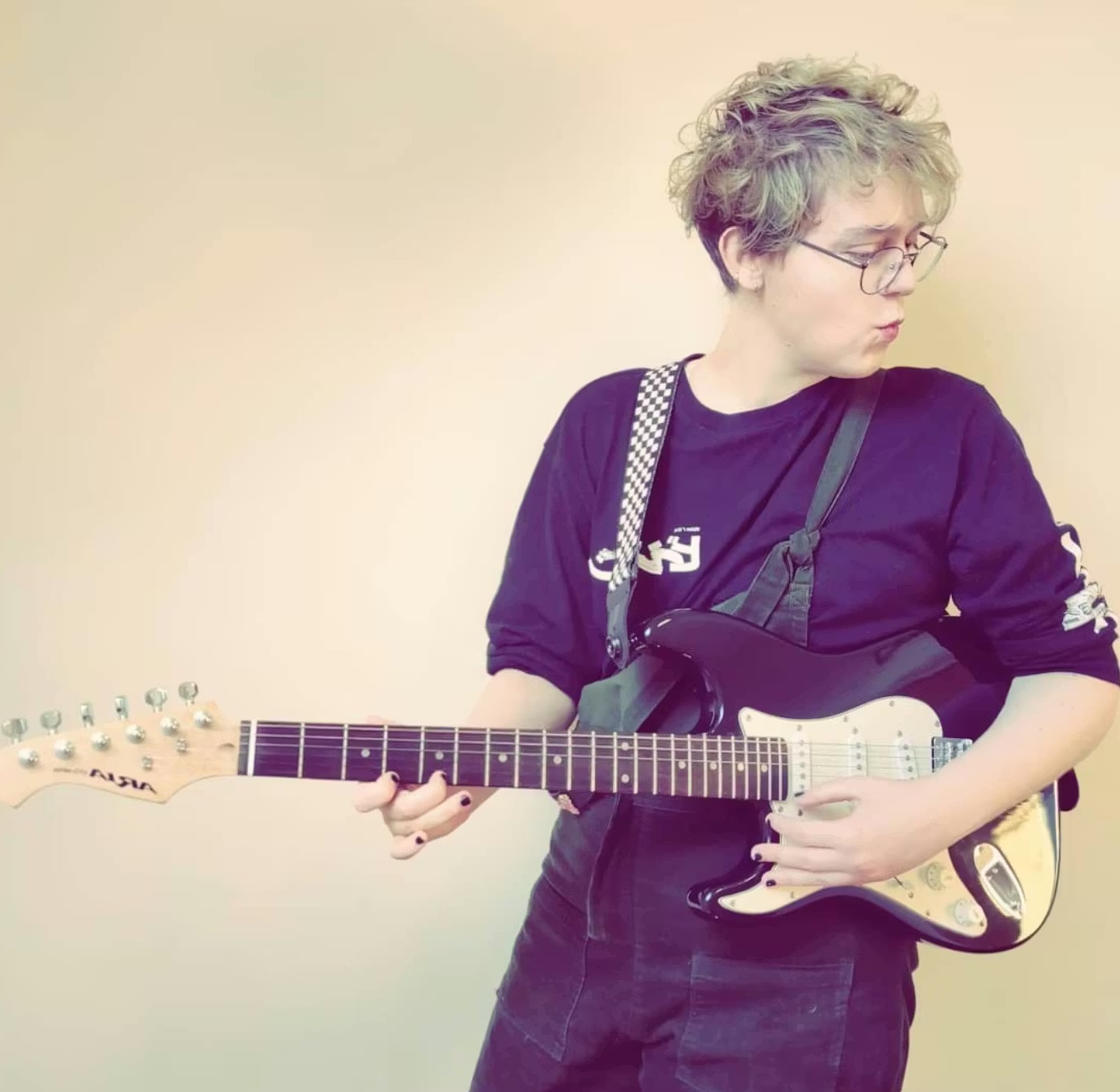 CHAINES (Cee Haines) stood holding a guitar and looking to their right.
