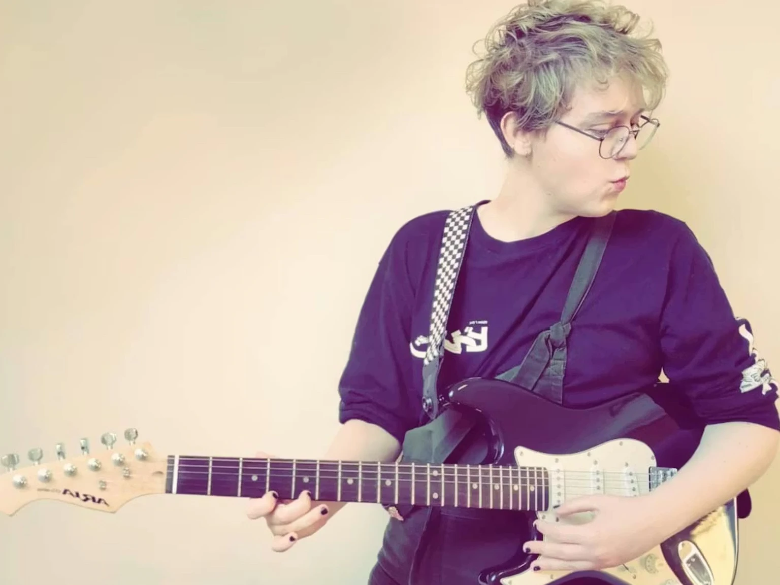 CHAINES (Cee Haines) stood holding a guitar and looking to their right.