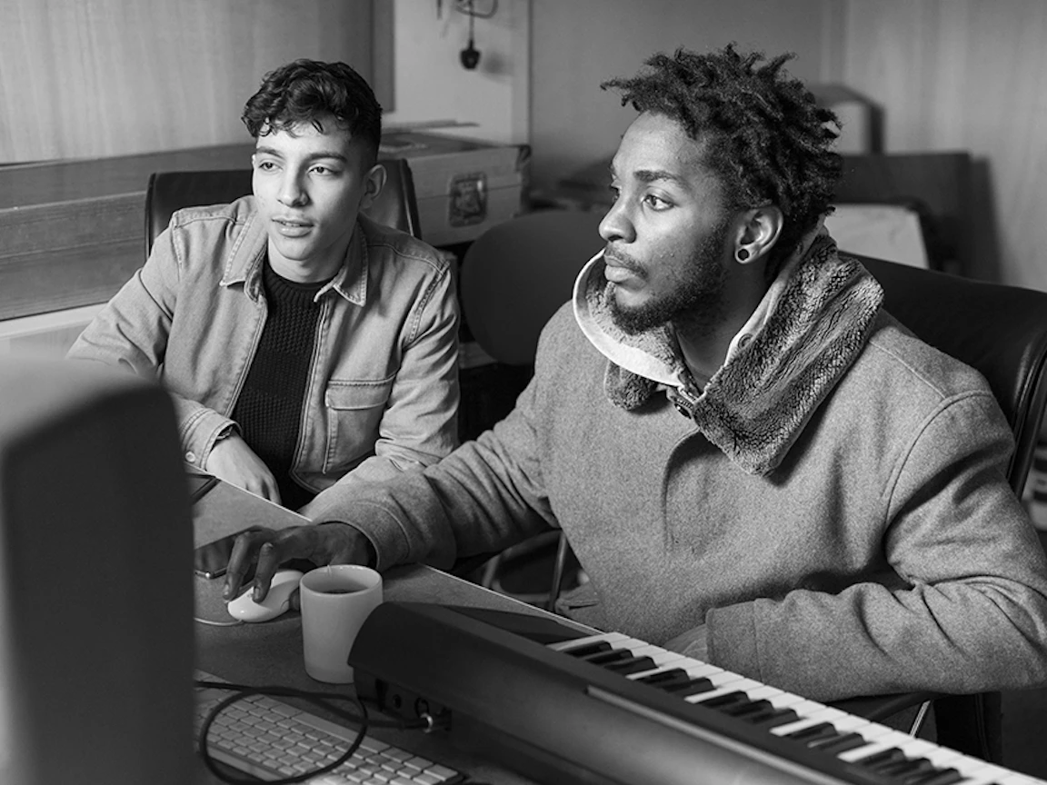 A young musician sat by a facilitator by a desk watching the screen.