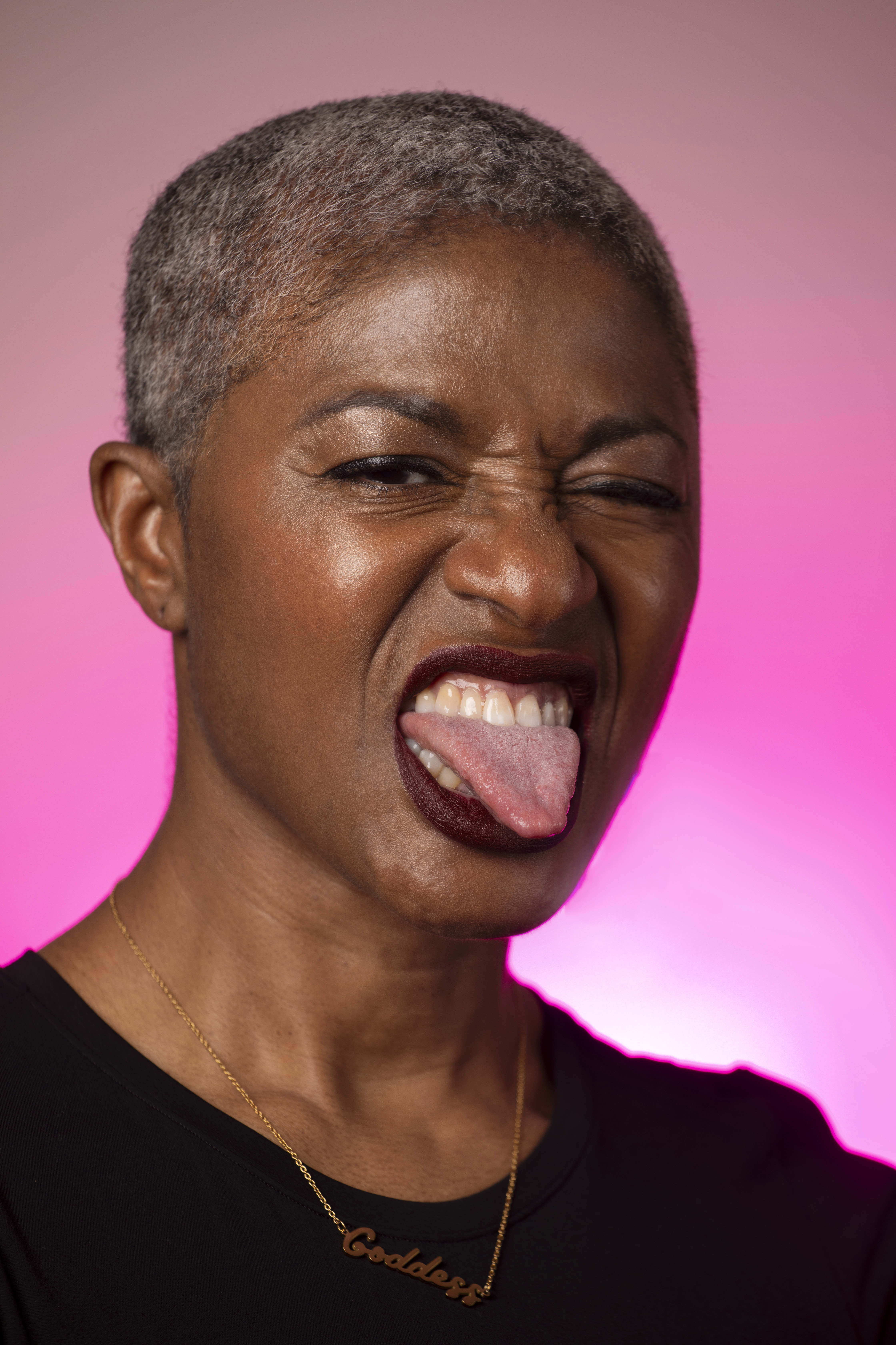 DJ Paulette with her tongue out on a background illuminated by a pink light.