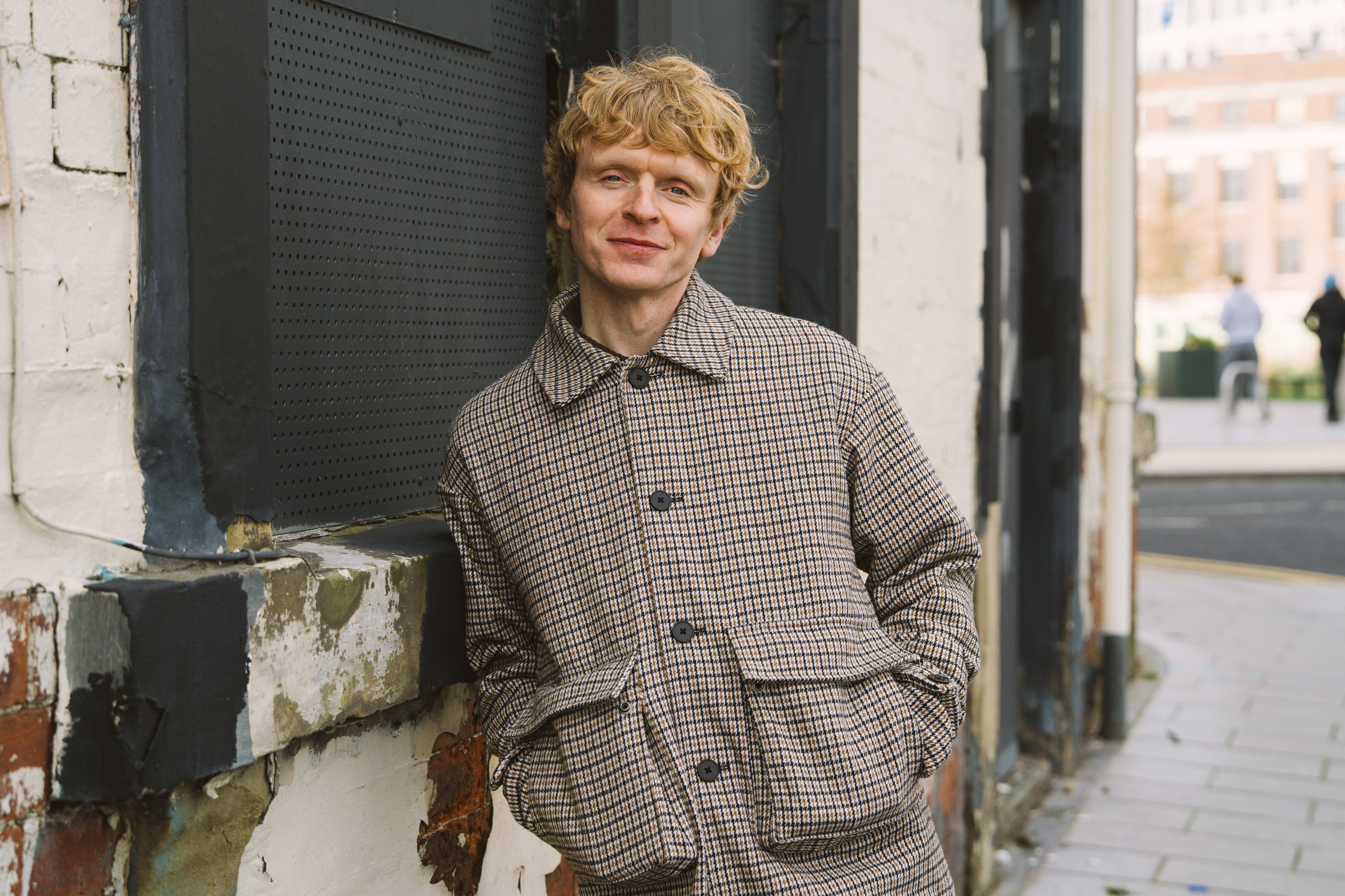 Scott Lewis looking to camera with his hands in his pockets leaning against a wall.