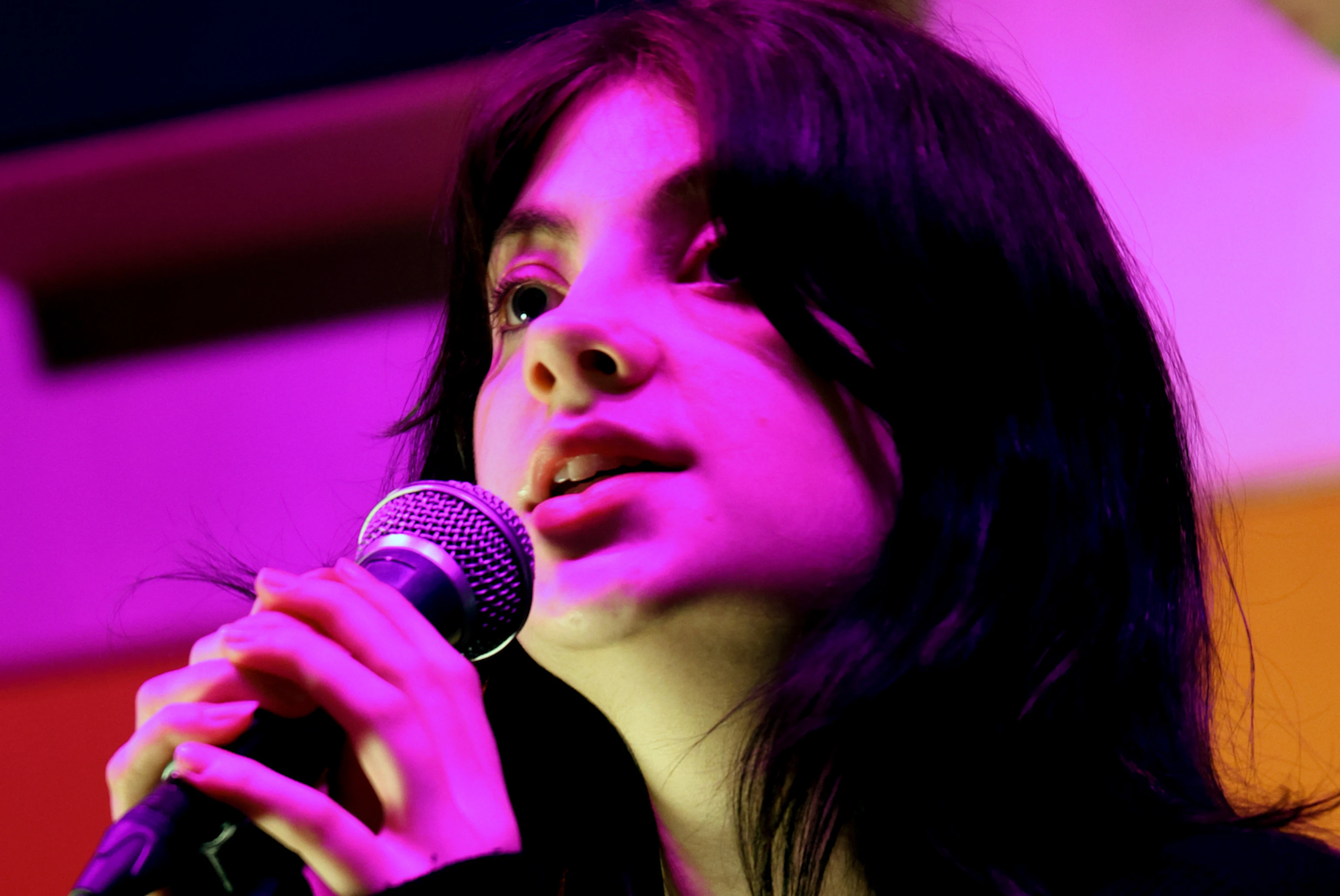 A close up of Jenni Orlopp singing into a microphone. She is illuminated by pink light.