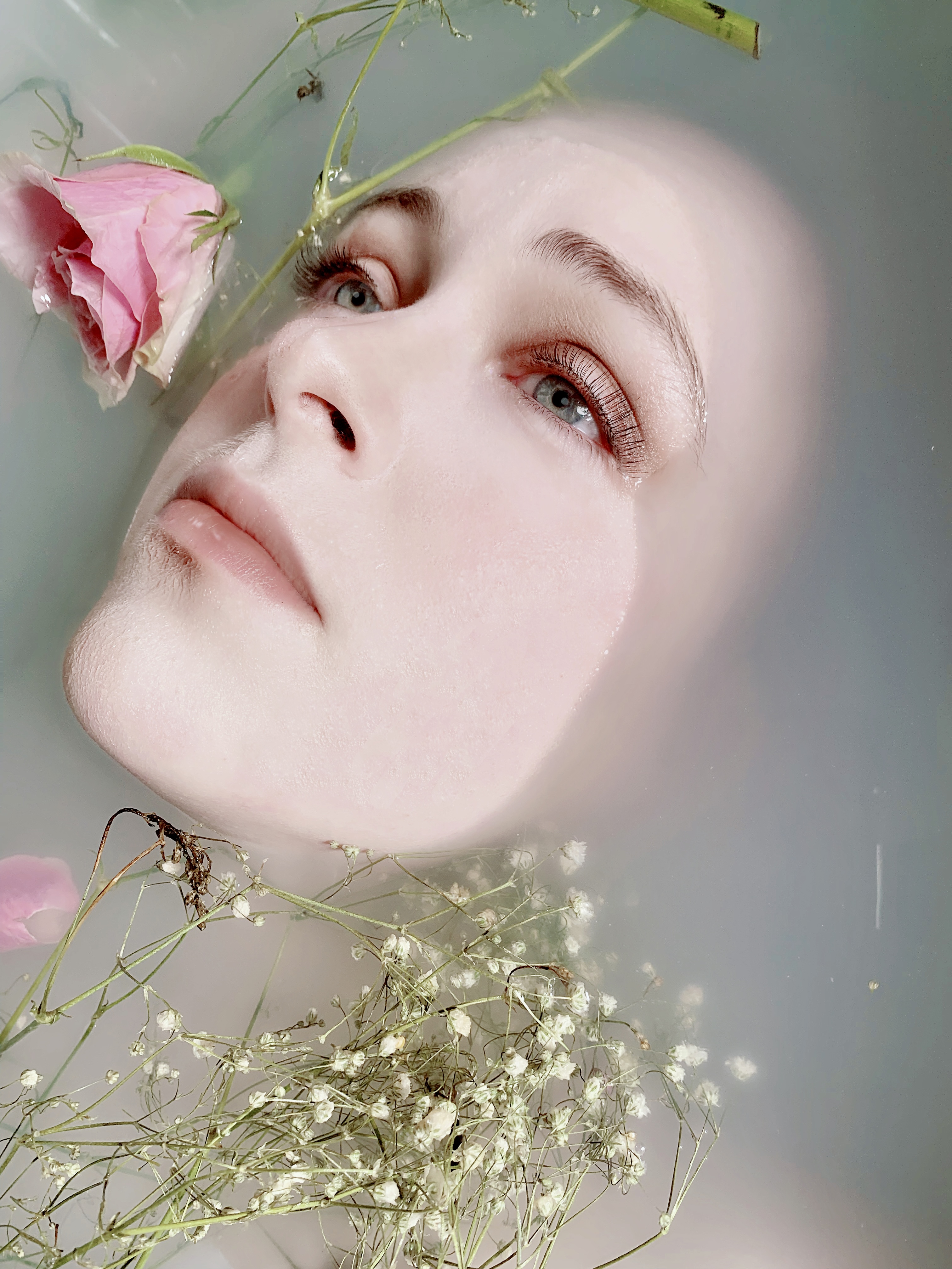 Charlotte Marlow's head is almost submerged in translucent water. Only their face is still visible. Surrounding their face are pink roses floating in the water.