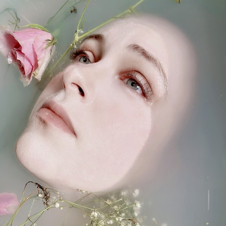 Charlotte Marlow's head is almost submerged in translucent water. Only their face is still visible. Surrounding their face are pink roses floating in the water.