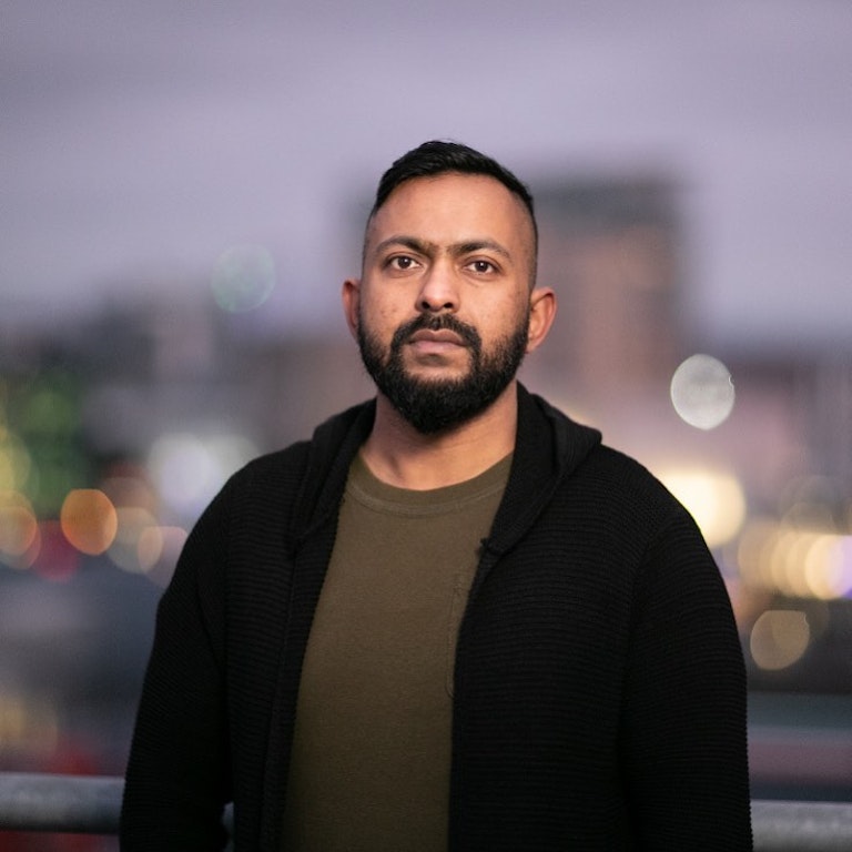 Sam Malik dressed in a black coat and a khaki t-shirt looking into the background on a faded background of a city skyline.