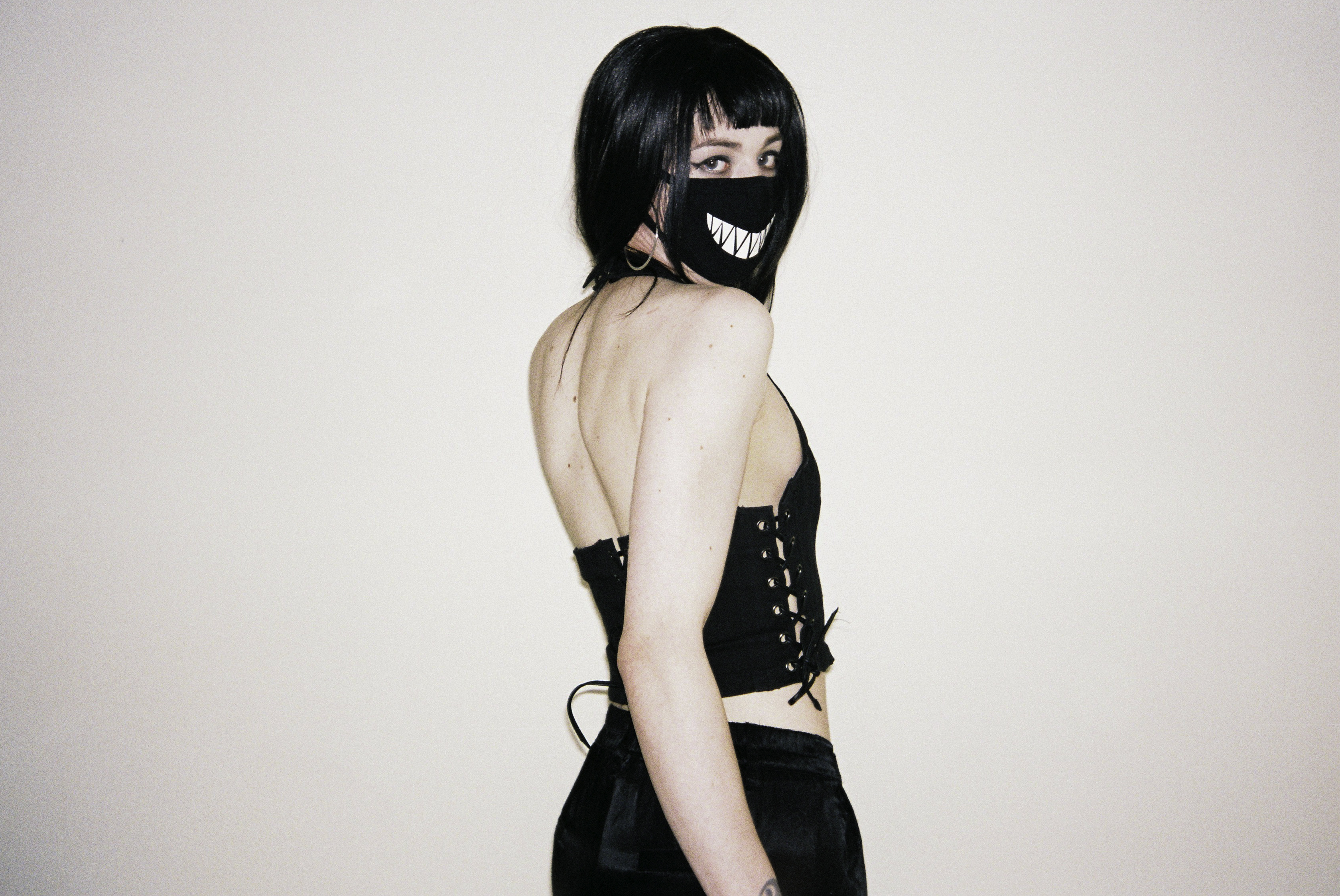 Syn on a white background with her body away from the camera and looking over her shoulder into the camera. She is wearing black attire, including a black face mask with white jagged teeth printed on the front.