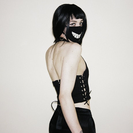 Syn on a white background with her body away from the camera and looking over her shoulder into the camera. She is wearing black attire, including a black face mask with white jagged teeth printed on the front.