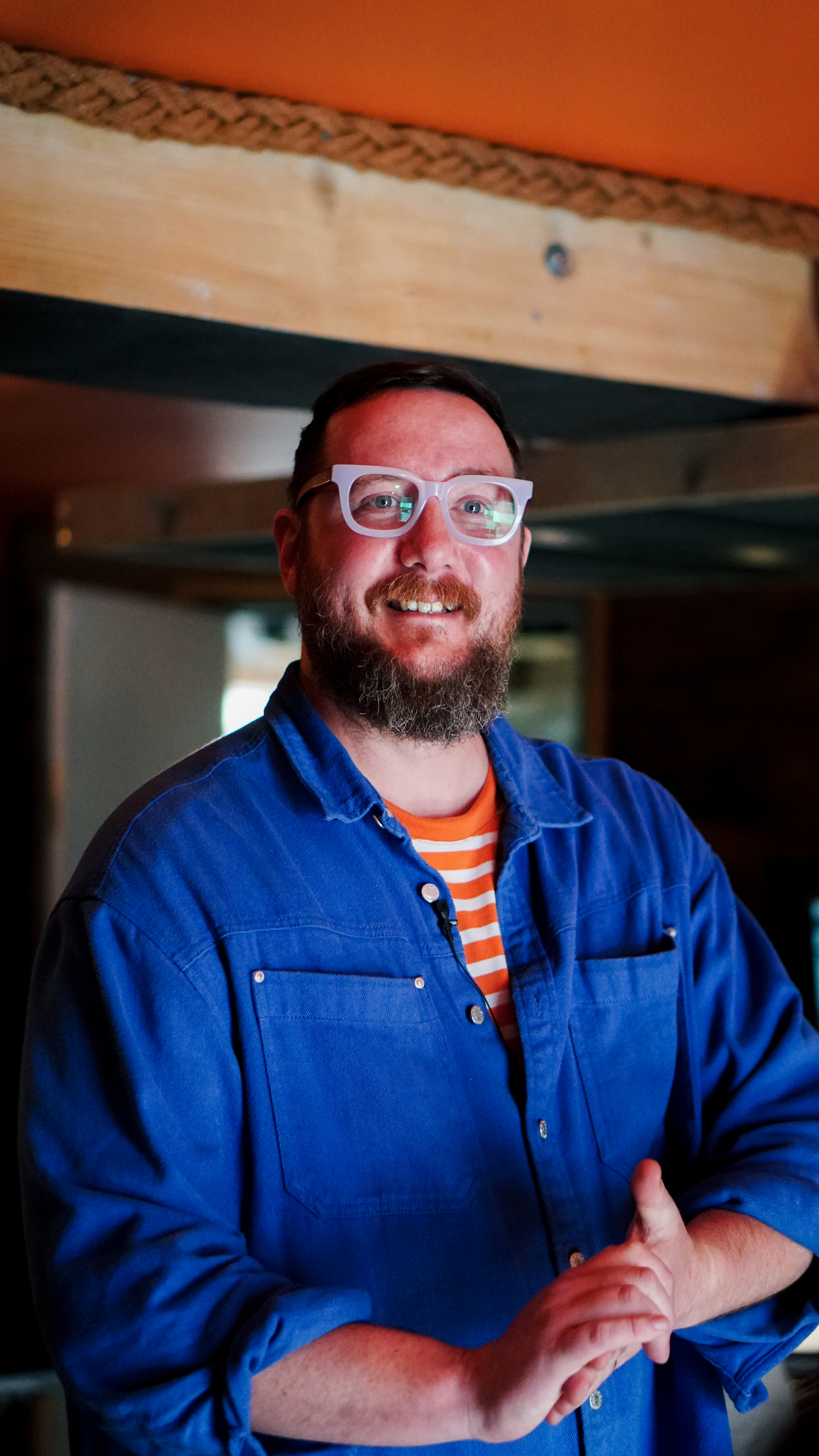 A photo of Phill Howley smiling broadly. He is wearing a bright blue shirt and wearing pale rimmed glasses.