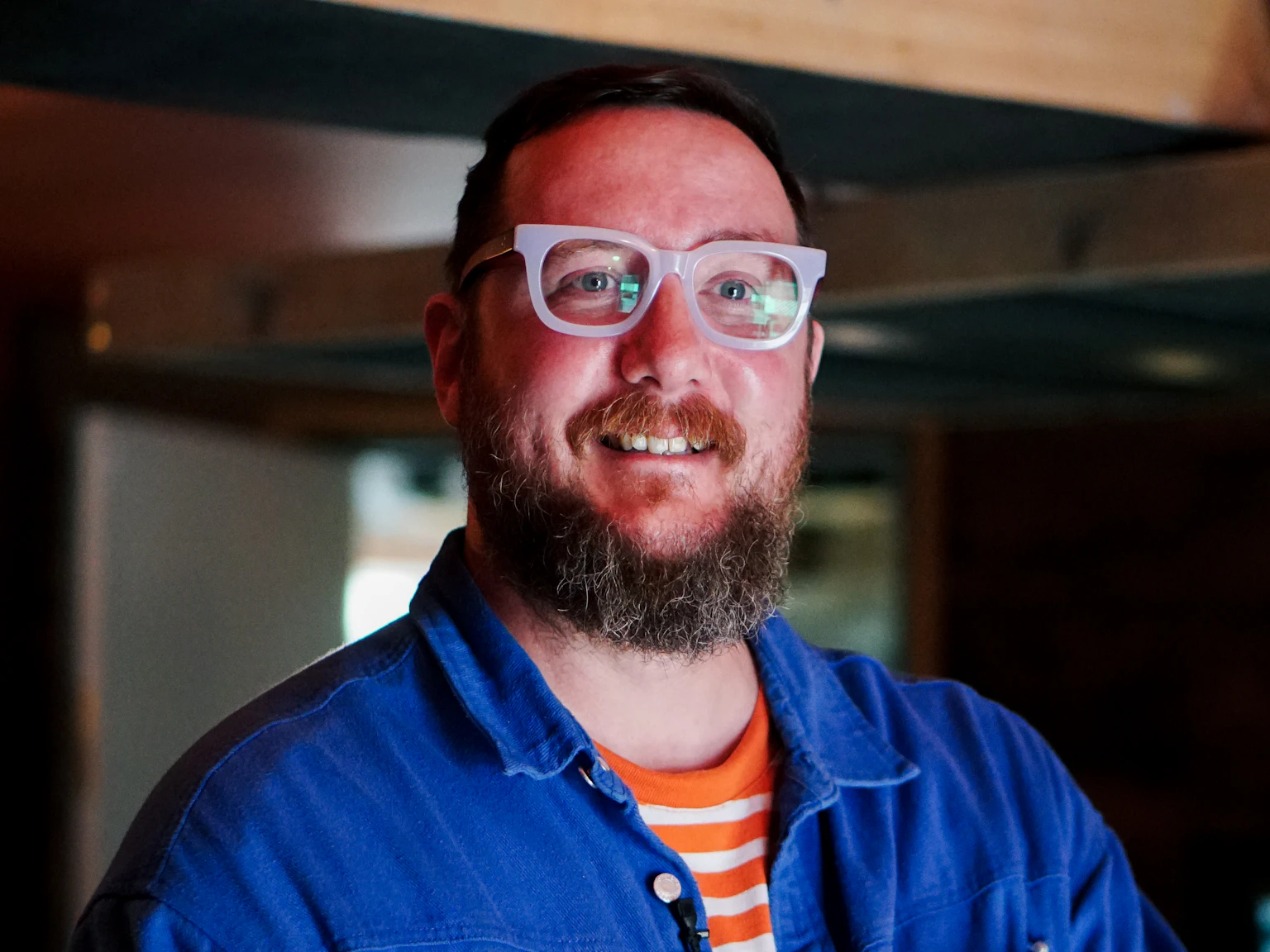 A photo of Phill Howley smiling broadly. He is wearing a bright blue shirt and wearing pale rimmed glasses.