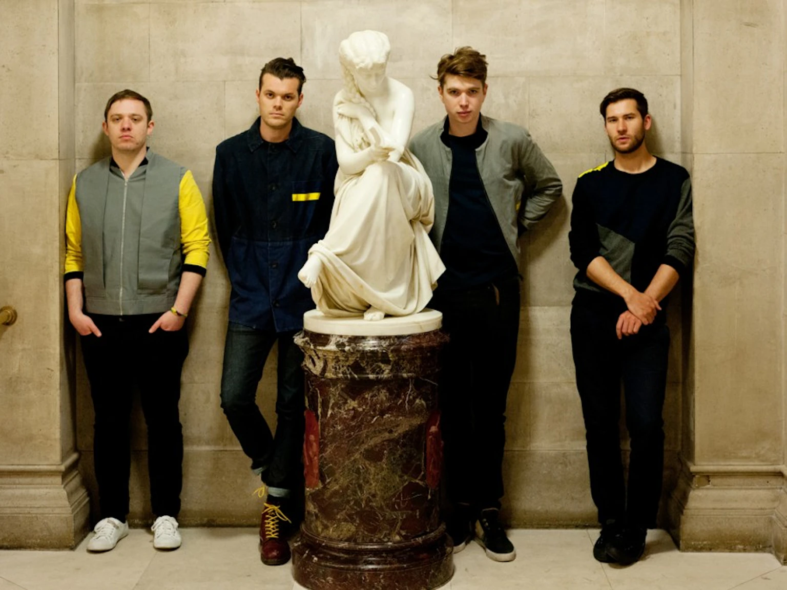 Everything Everything in a row stood against a stone wall looking at the camera with a marble statue in the middle of them.