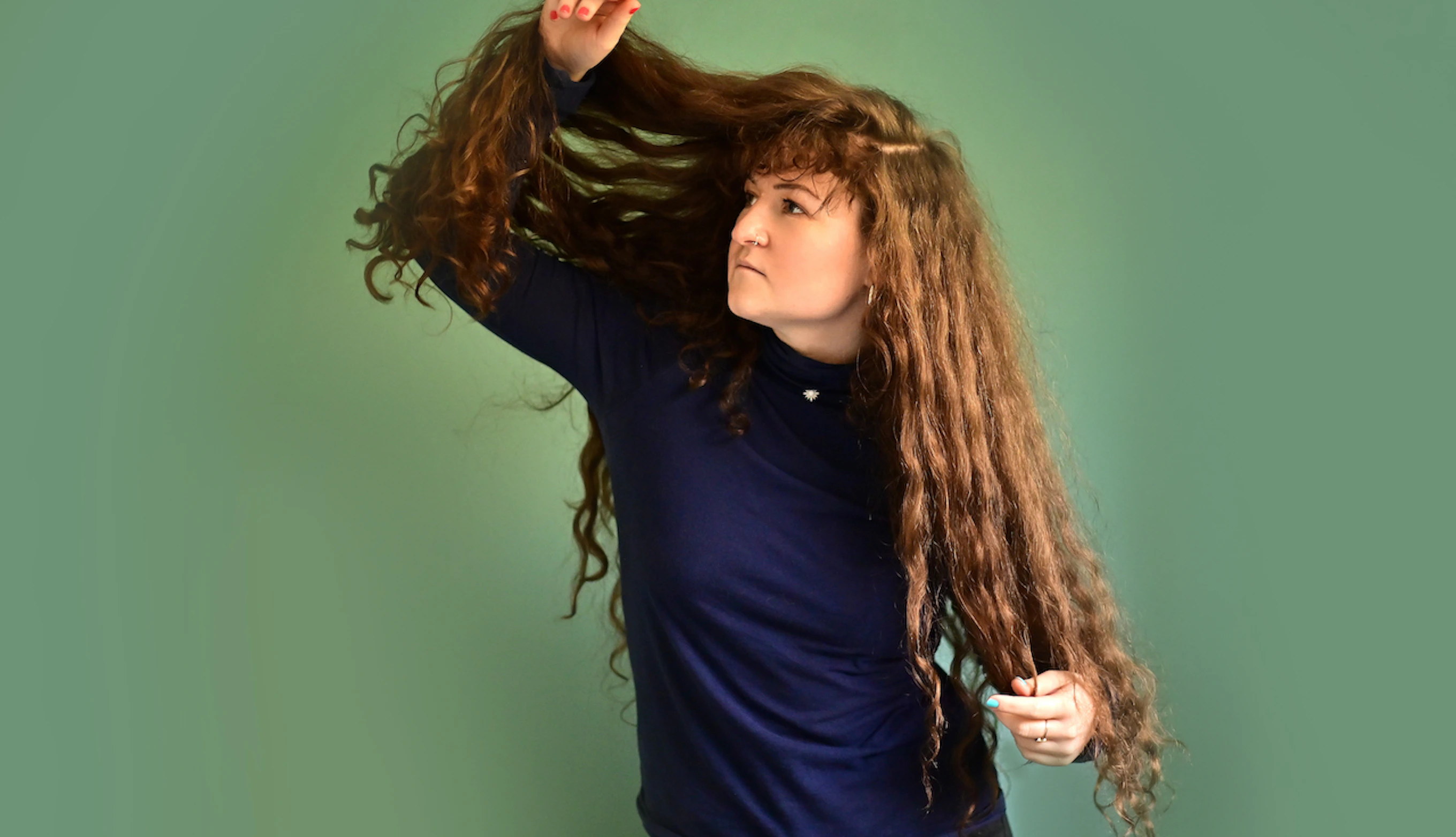 IORA stands against a green background looking to one side. She is lifting one side of her long wavy hair up with her arm.