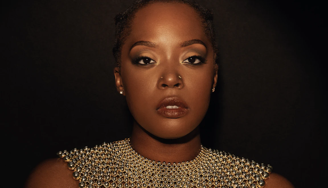 Karen Nyame KG dressed in black with a loose gold choker necklace made from beads, and a bracelet. Both hands are holding her arms as she stares into the camera.