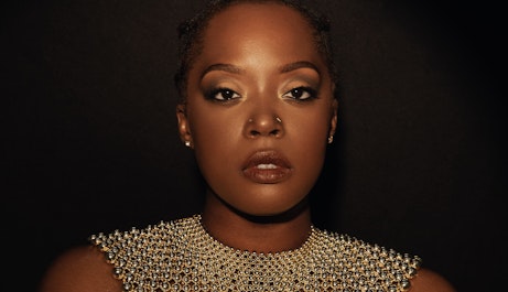 Karen Nyame KG dressed in black with a loose gold choker necklace made from beads, and a bracelet. Both hands are holding her arms as she stares into the camera.
