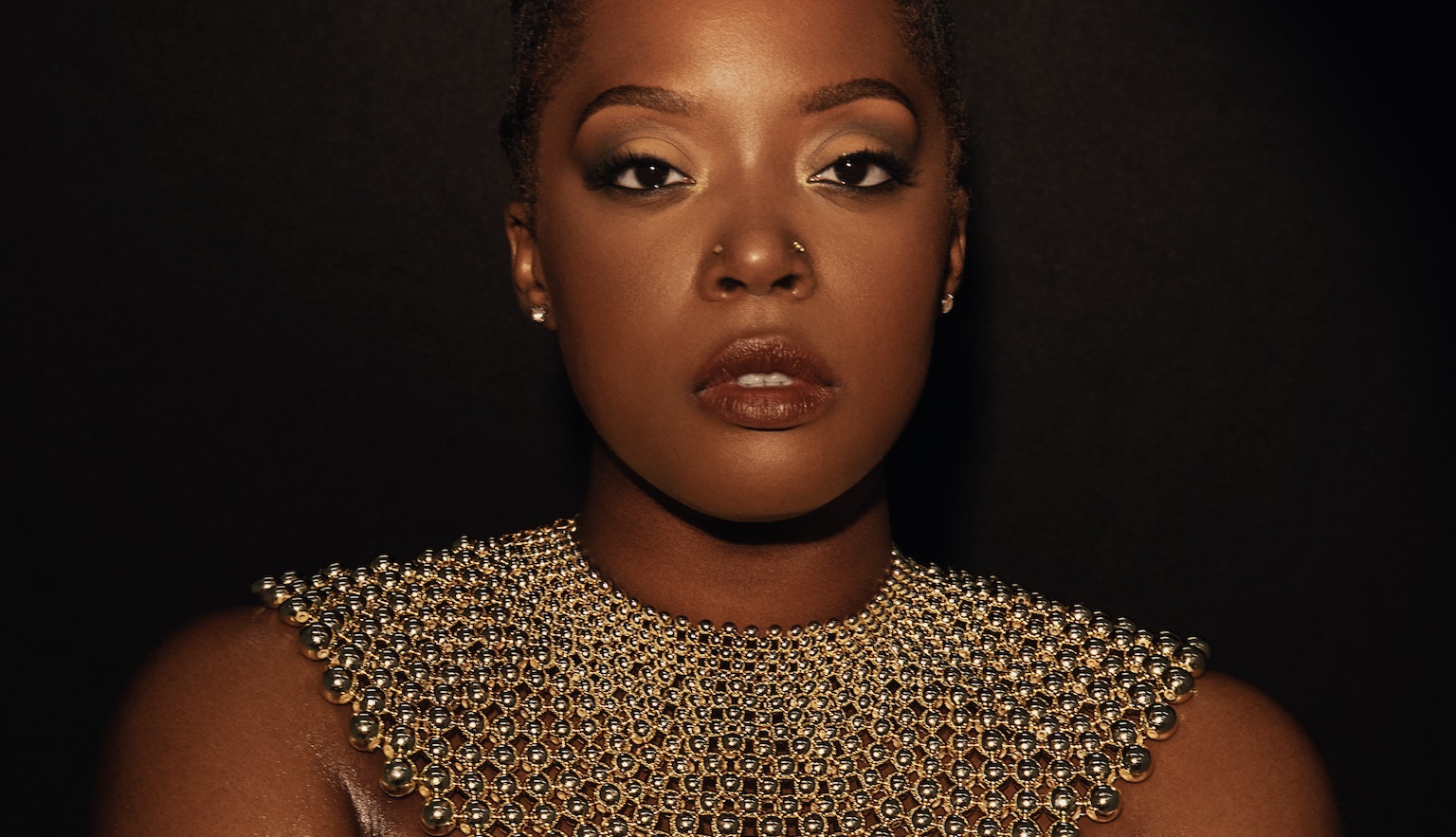 Karen Nyame KG dressed in black with a loose gold choker necklace made from beads, and a bracelet. Both hands are holding her arms as she stares into the camera.