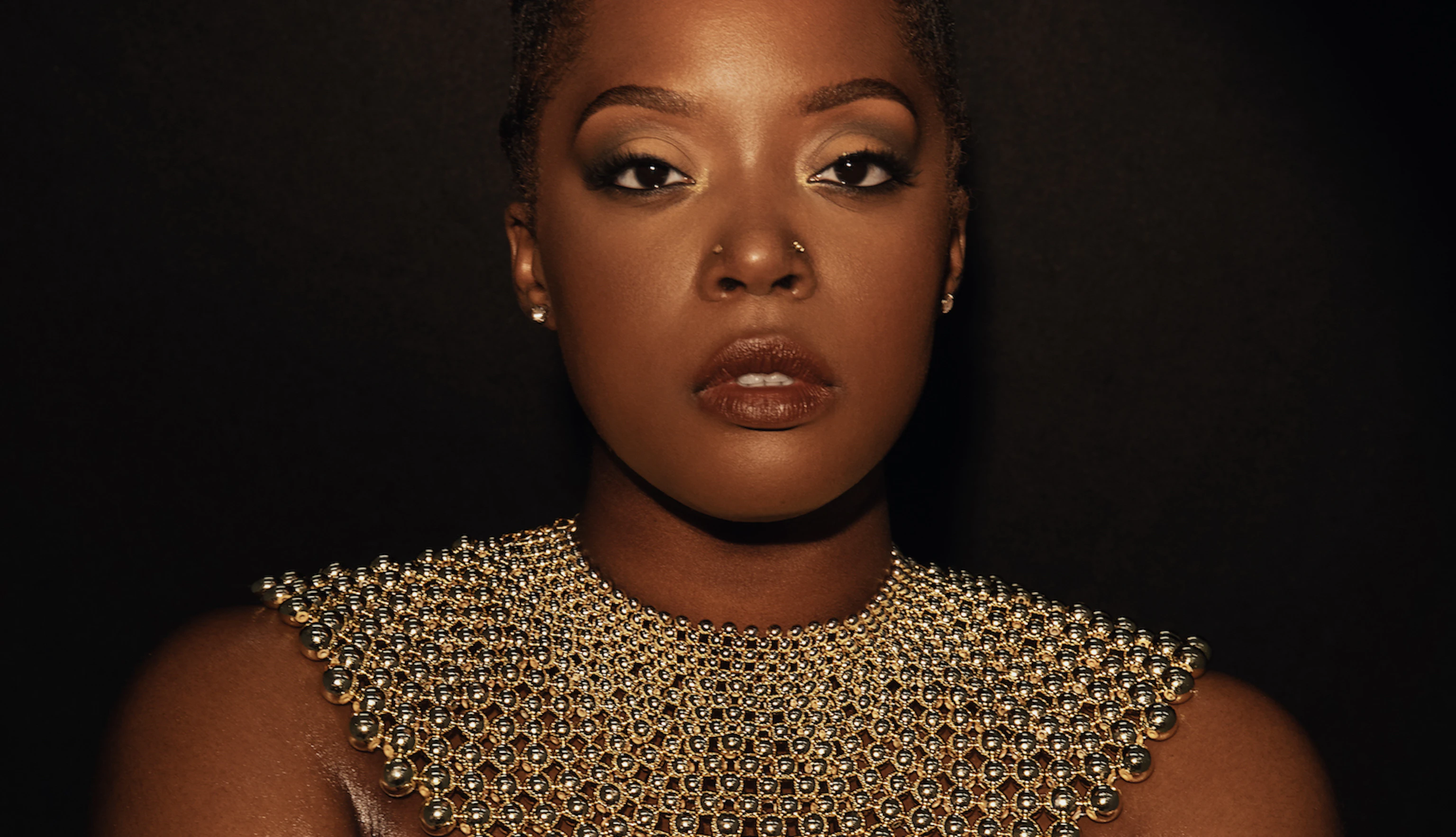 Karen Nyame KG dressed in black with a loose gold choker necklace made from beads, and a bracelet. Both hands are holding her arms as she stares into the camera.