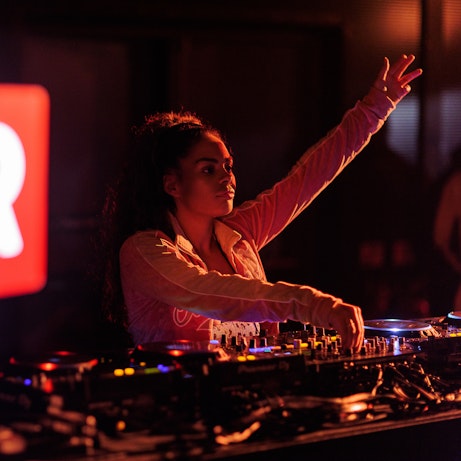 DJ Chaise with one hand in the air, the other adjusting a knob on the decks. The Brighter Sound lightbox is encroaching on the left hand side slightly blurred.