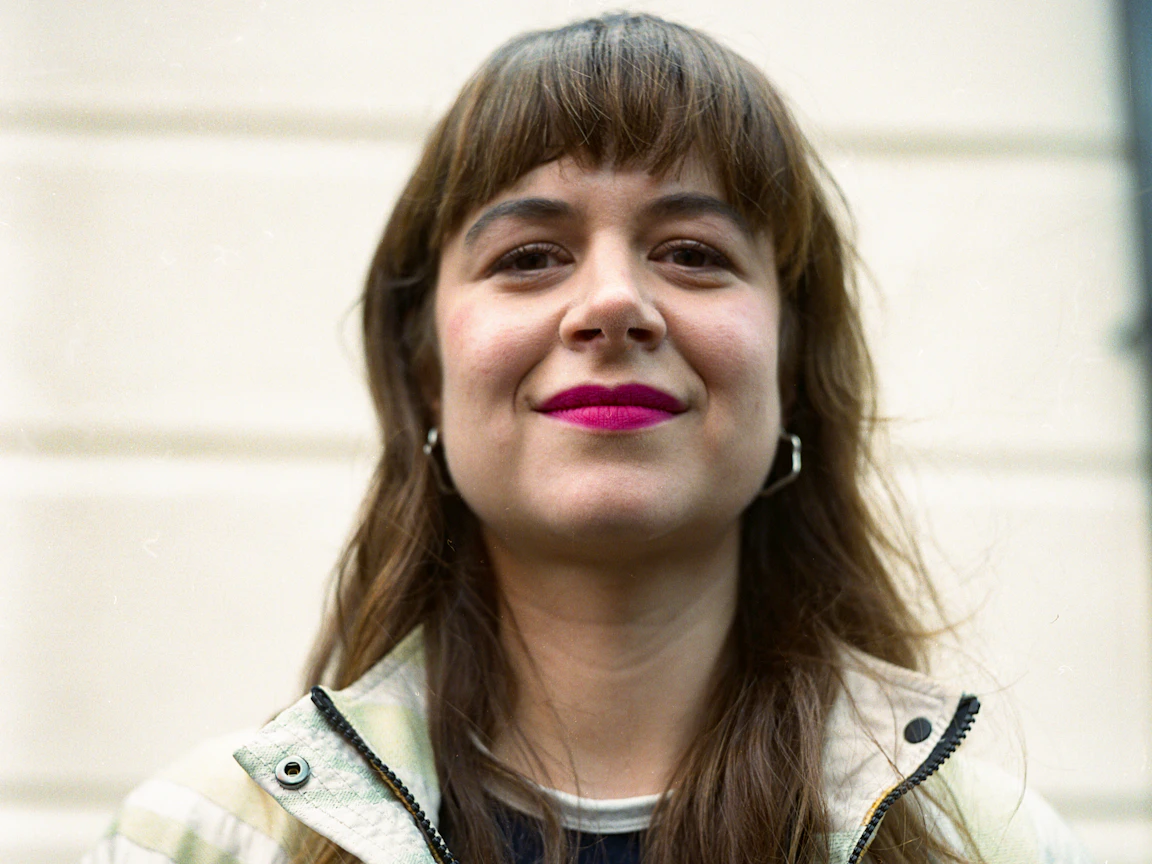 Rebecca is a white woman in her mid 30s. She has dark mid-length hair with a fringe. Her lipstick is a very bright shade of pink.