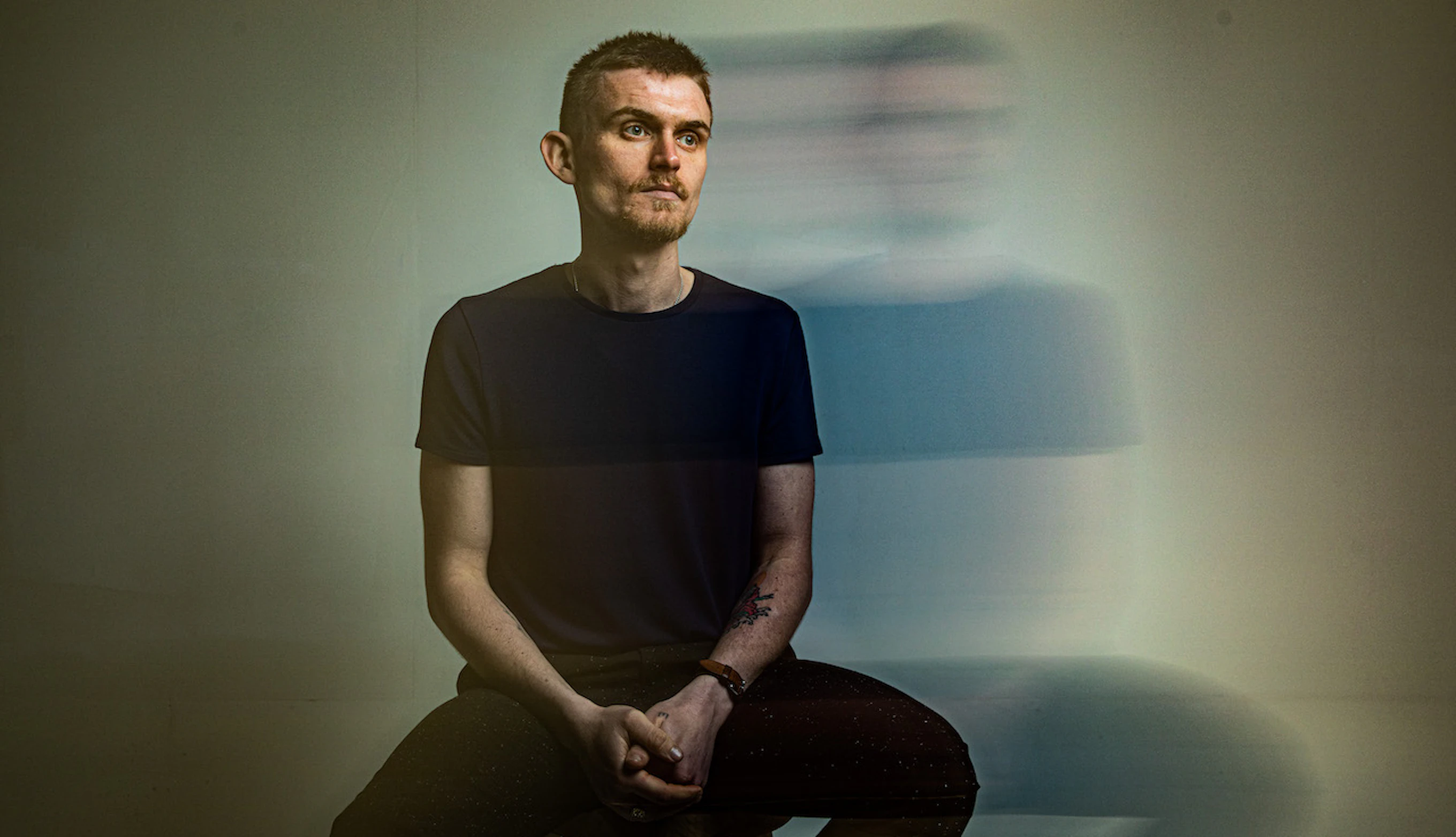 Harry Heart is sitting on a stool, looking into the distance. The background is plain although the camera has created an effect in which his outline is repeated in a blur next to him.