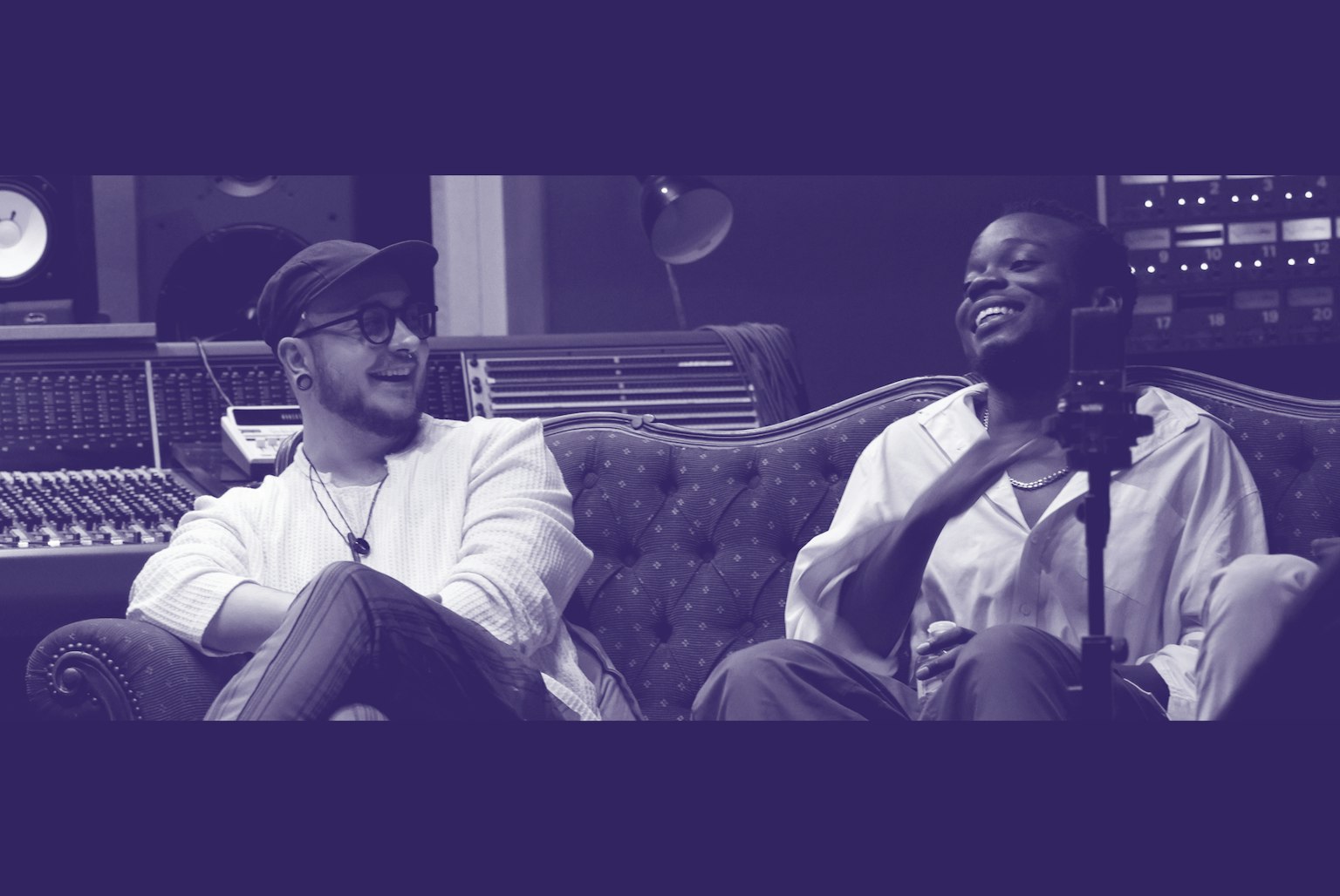 Two people sat on a couch. One is looking at the other and both are laughing. The photo is tinted navy blue and white.