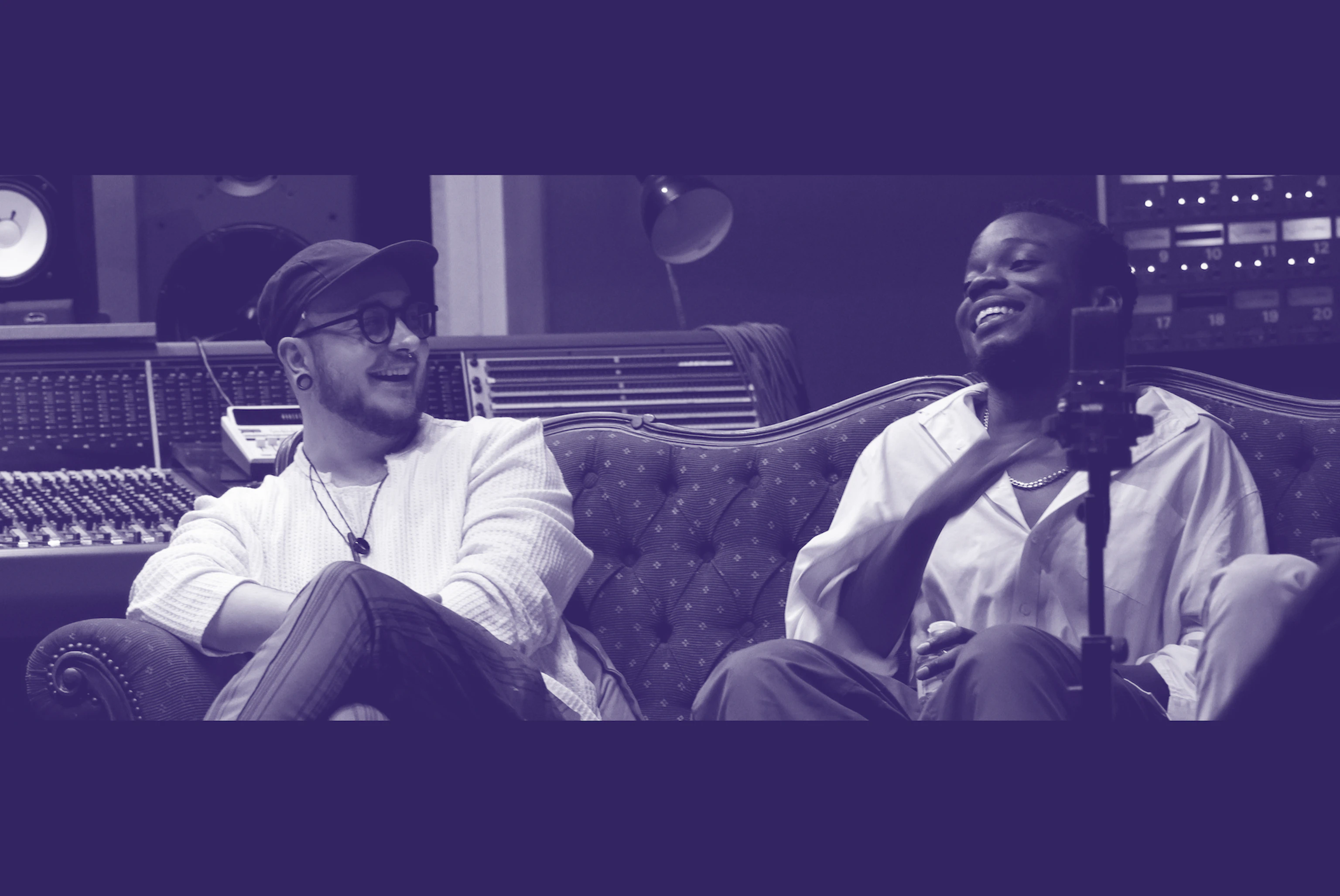 Two people sat on a couch. One is looking at the other and both are laughing. The photo is tinted navy blue and white.