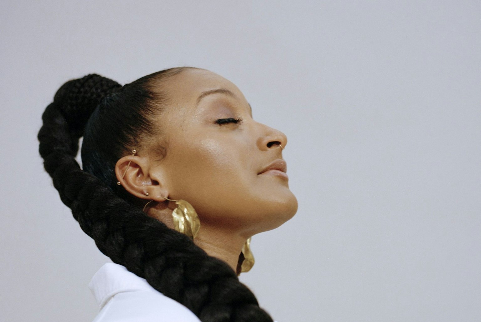 The artist Nubya Garcia has their eyes closed and head tilted upwards. Nubya is wearing a crisp white shirt and gold hoop earrings. Nubya's hair is plaited and cascades down over their shoulder. 