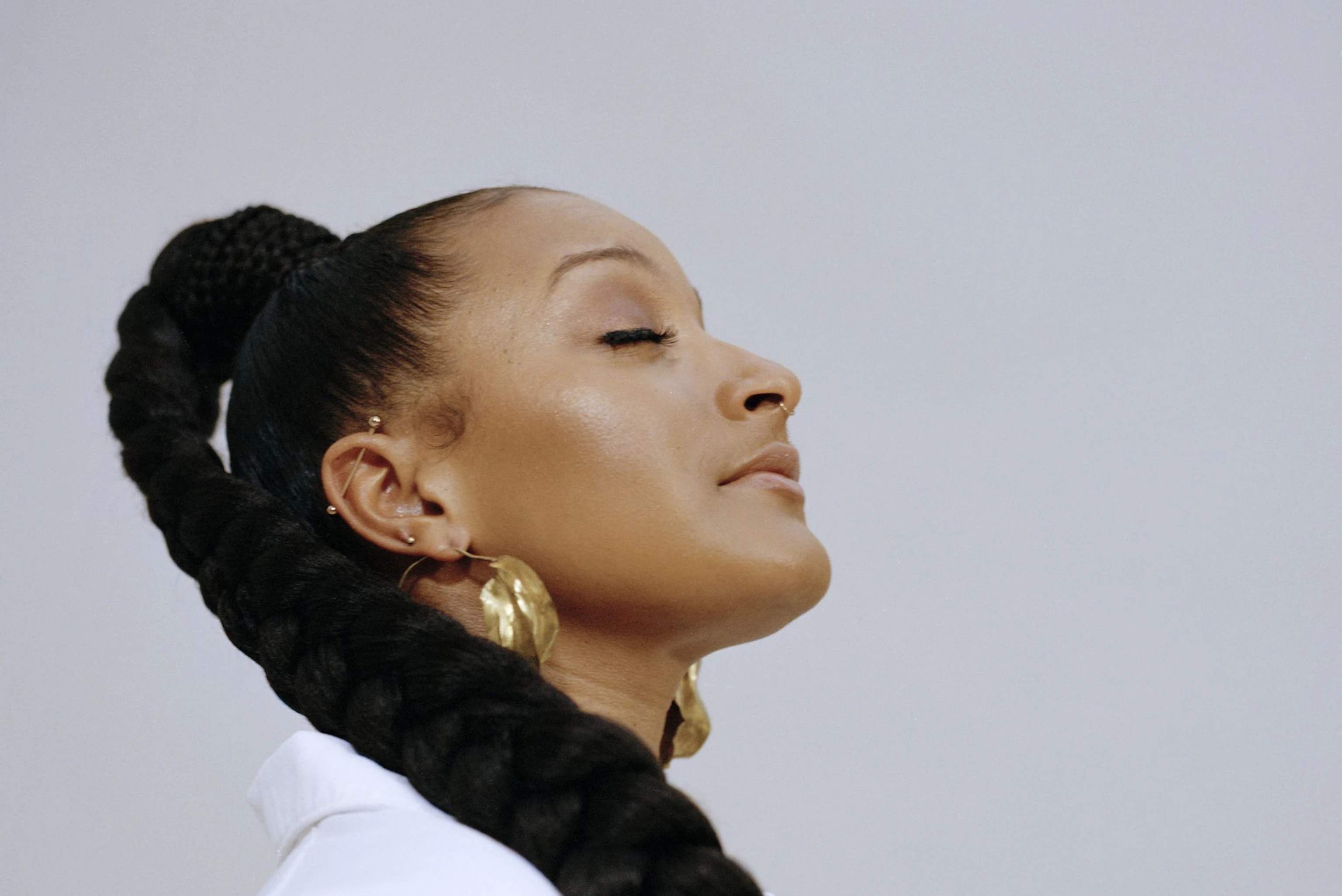 The artist Nubya Garcia has their eyes closed and head tilted upwards. Nubya is wearing a crisp white shirt and gold hoop earrings. Nubya's hair is plaited and cascades down over their shoulder.