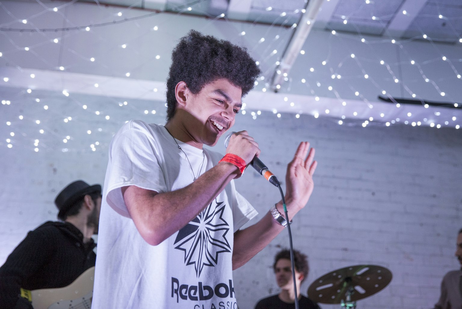 A young artist stands on stage performing with a microphone in hand. In the background is a band and some twinkling fairy lights.