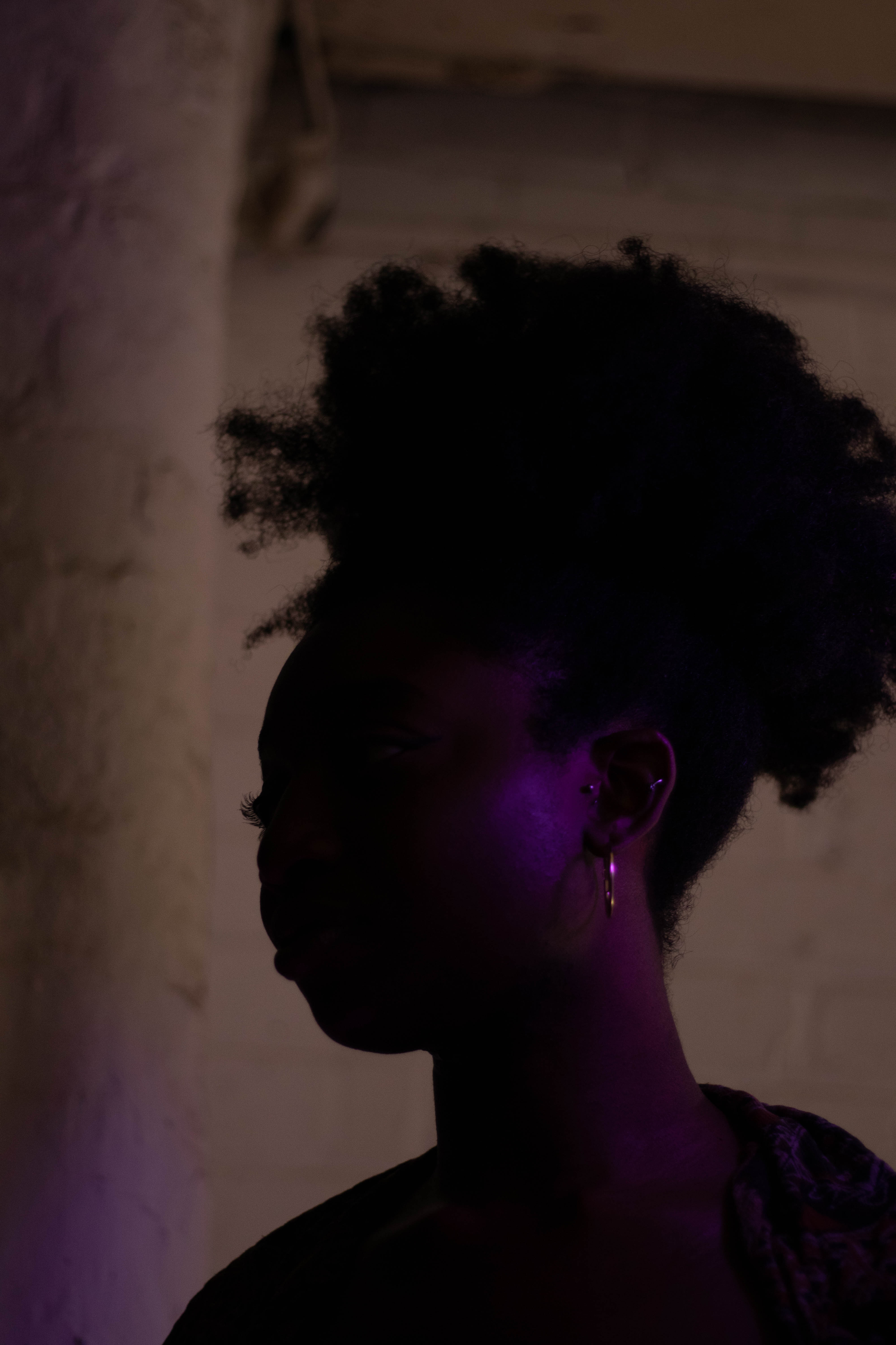 SCAPA in a dark space. Her face is hidden in shadow, the side of her head slightly illuminated by a purple light.