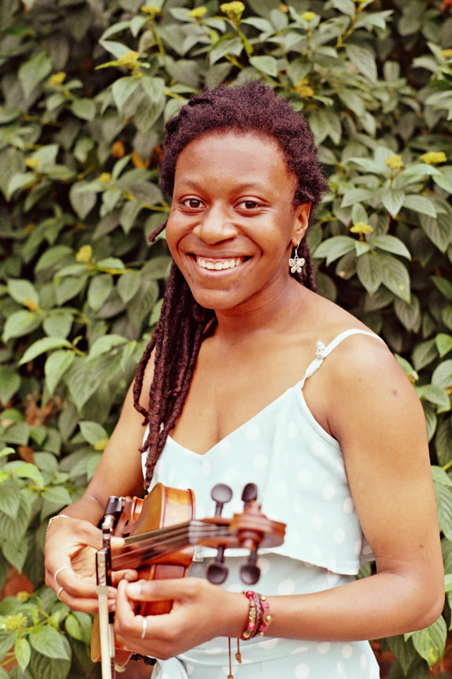 Sam Brown smiling into the camera and holding her violin on a background of green leaves.