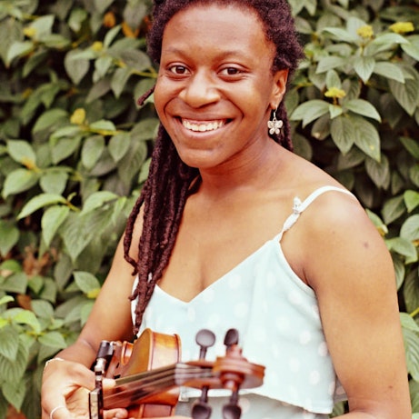 Sam Brown smiling into the camera and holding her violin on a background of green leaves.