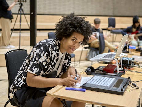 A music practitioner is looking up from writing on a desk, and smiling to the camera. They are surrounded by music equipment and young musicians can be seen in the background.