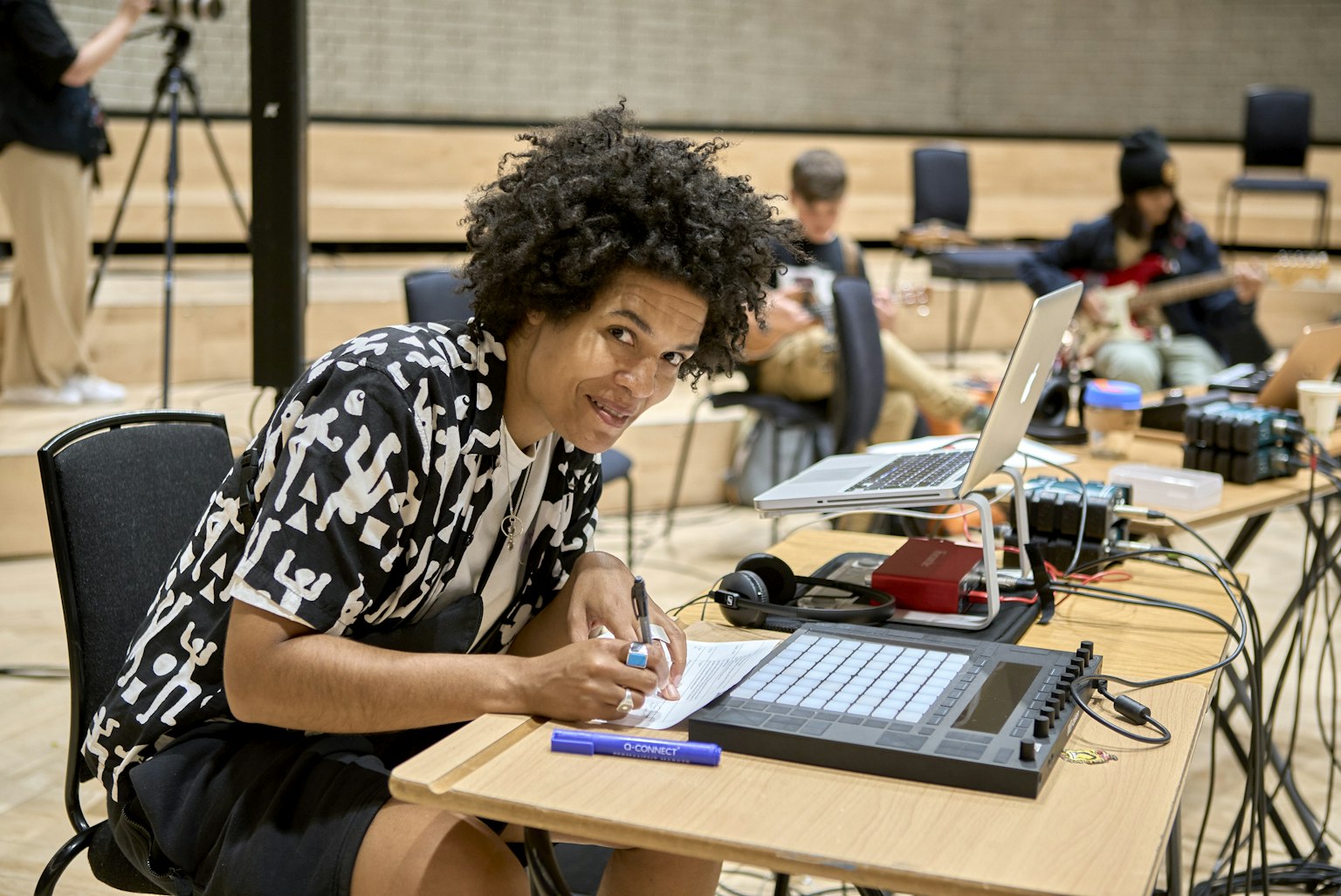 A music practitioner is looking up from writing on a desk, and smiling to the camera. They are surrounded by music equipment and young musicians can be seen in the background.