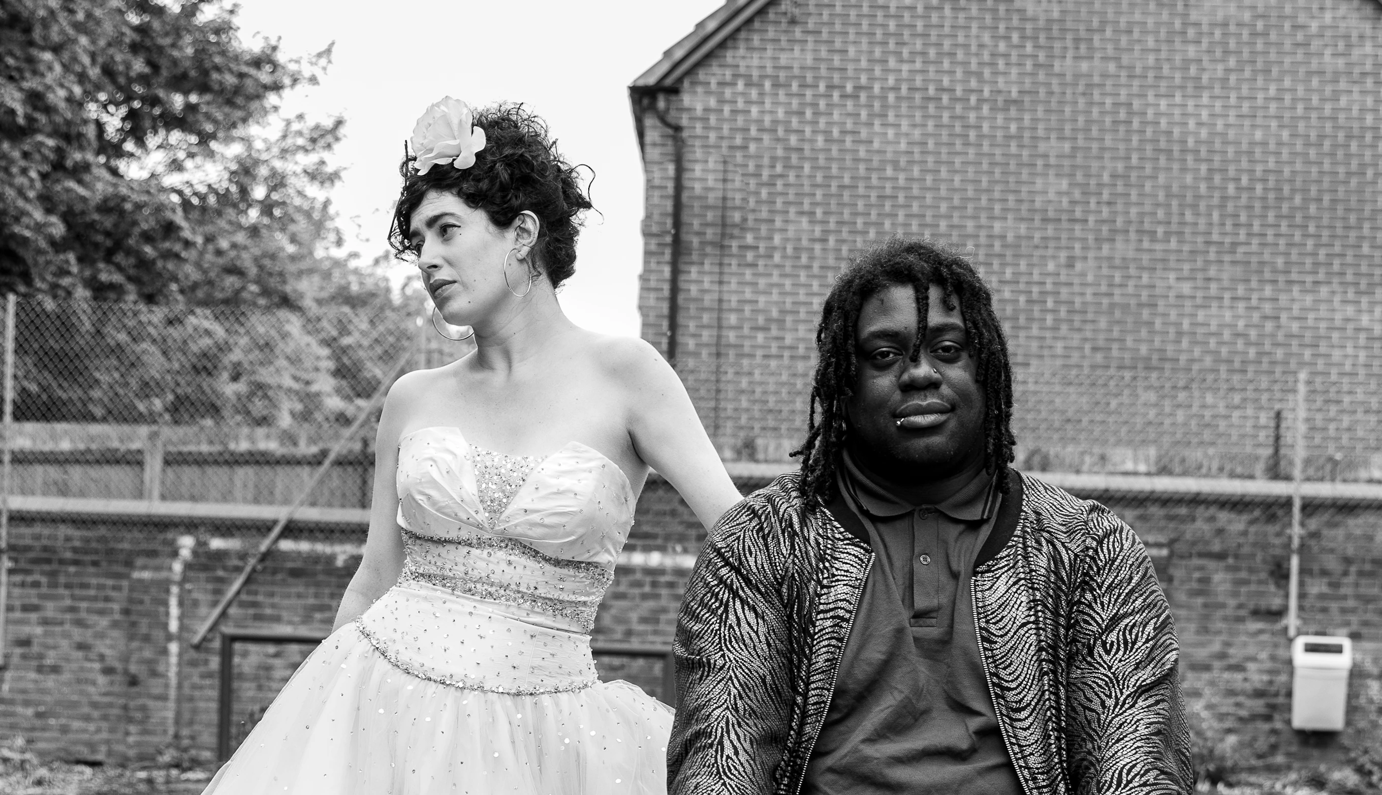 A black and white image of the two members of Dirty Freud. Ruby (to the left) is looking to one side. Danni (to the right) is looking directly at the camera. They are standing outside with a house and trees behind them.