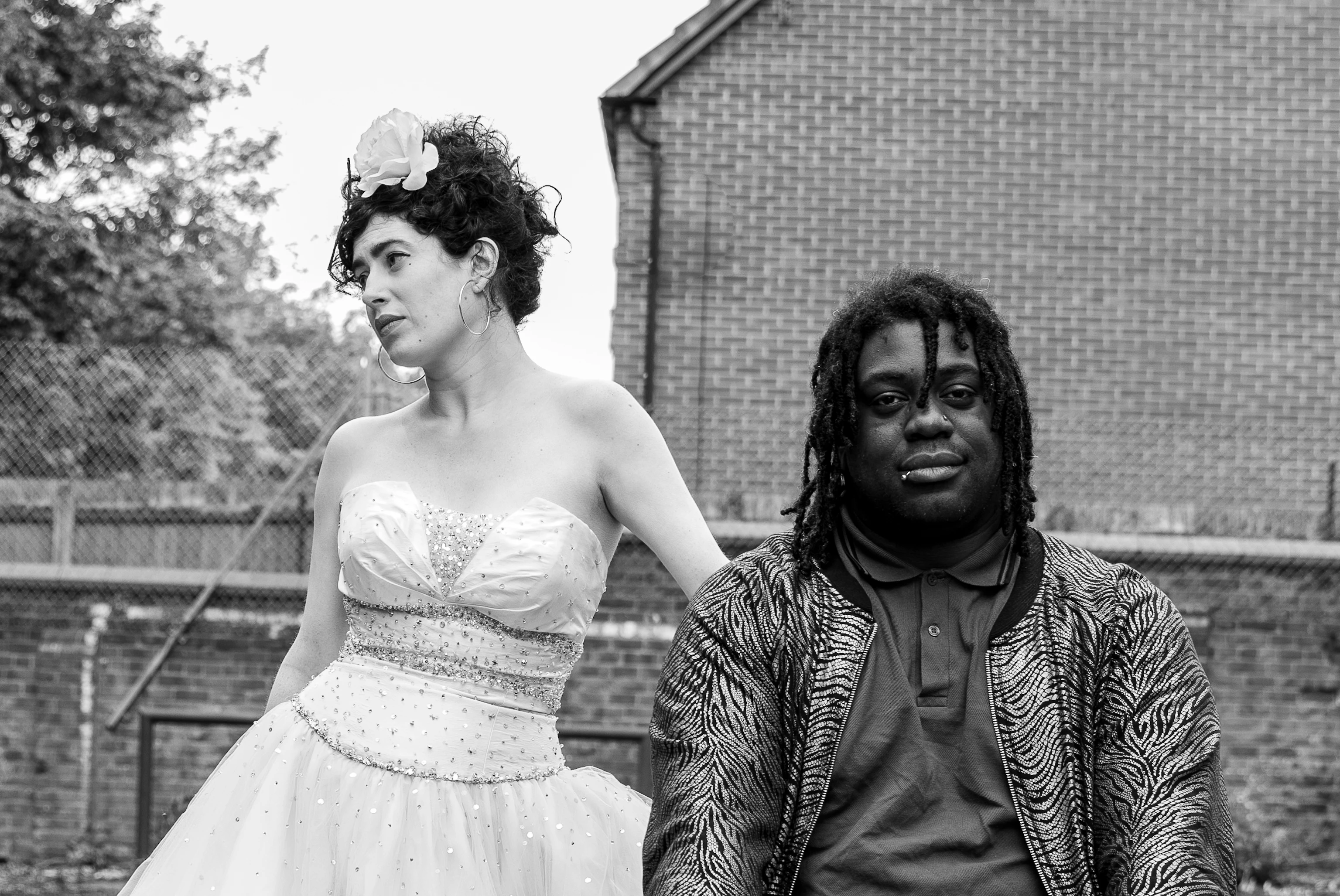 A black and white image of the two members of Dirty Freud. Ruby (to the left) is looking to one side. Danni (to the right) is looking directly at the camera. They are standing outside with a house and trees behind them.