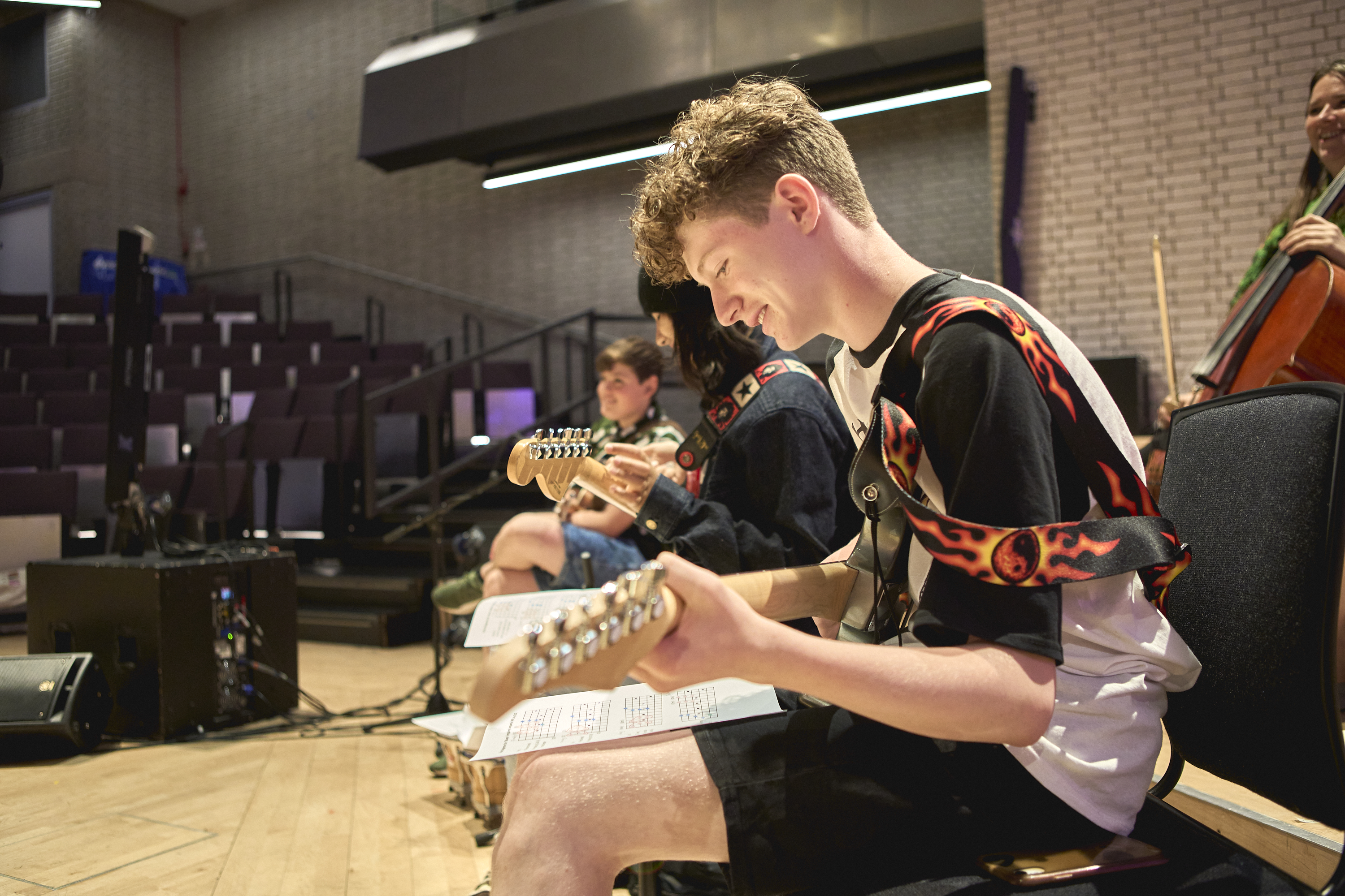 A side view of three young musicians sat in a row playing guitars. The one closest to the camera is smiling and looking down at sheet music on their lap.