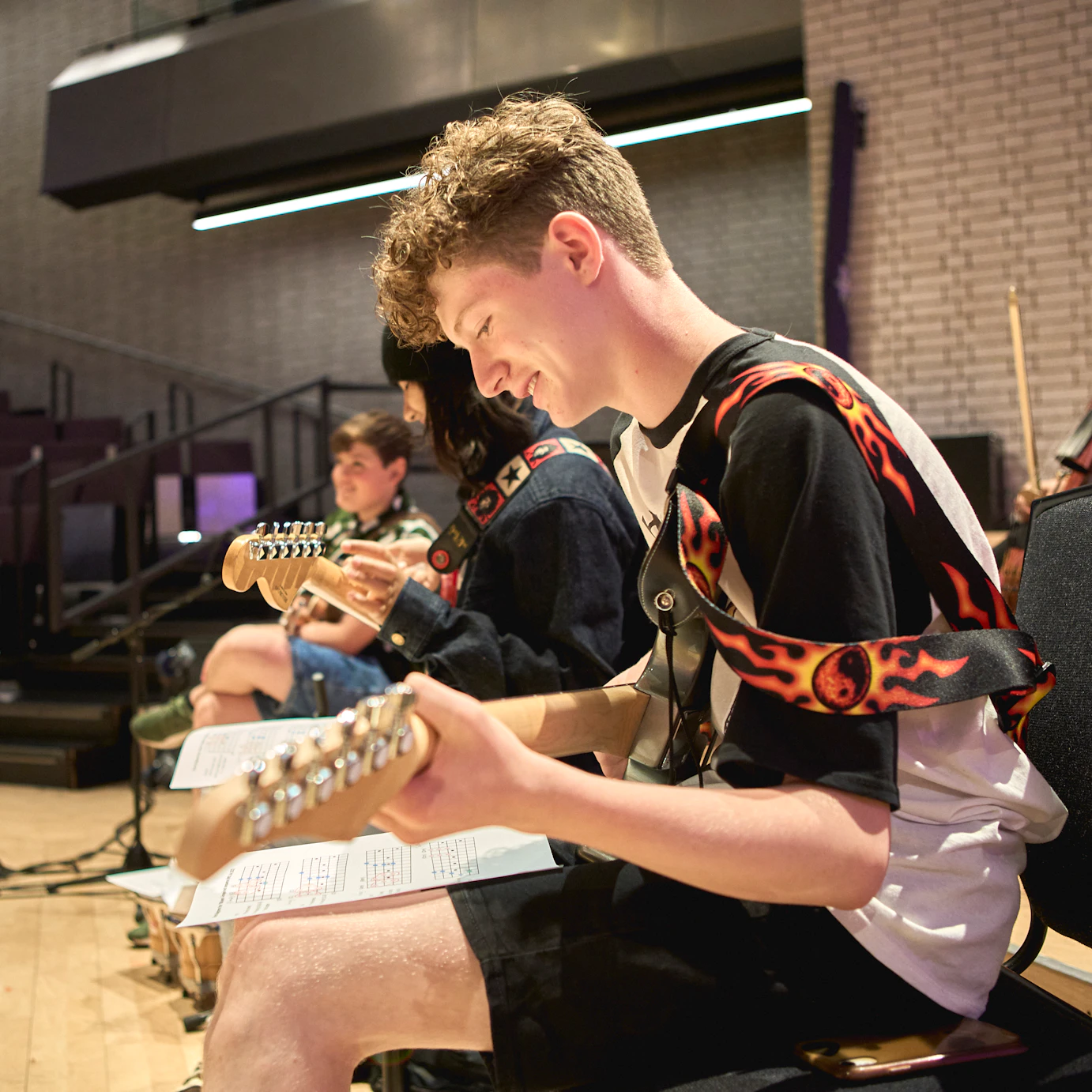 A side view of three young musicians sat in a row playing guitars. The one closest to the camera is smiling and looking down at sheet music on their lap.